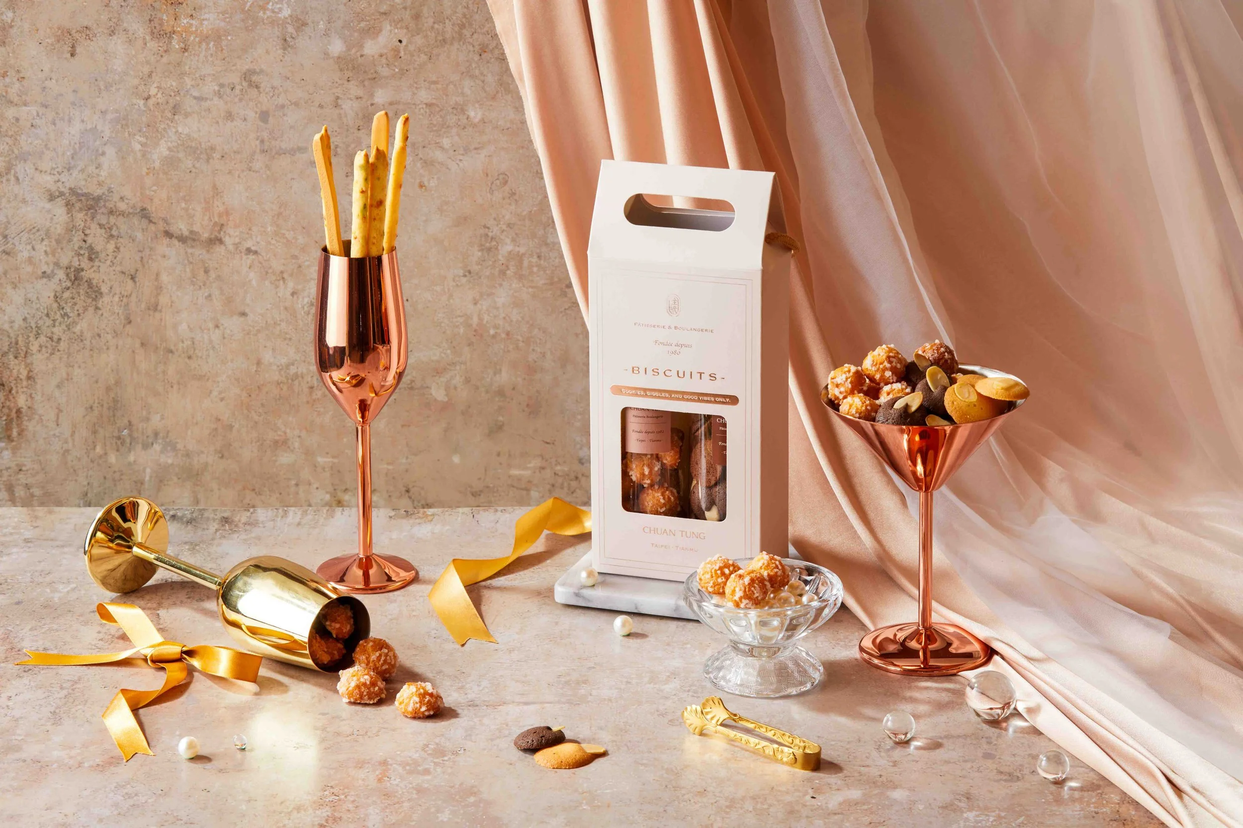 Decorative display of assorted cookies and biscuits in glass bowls and boxes, with gold and pink accents, on a marble surface against a textured wall and pink satin fabric backdrop.