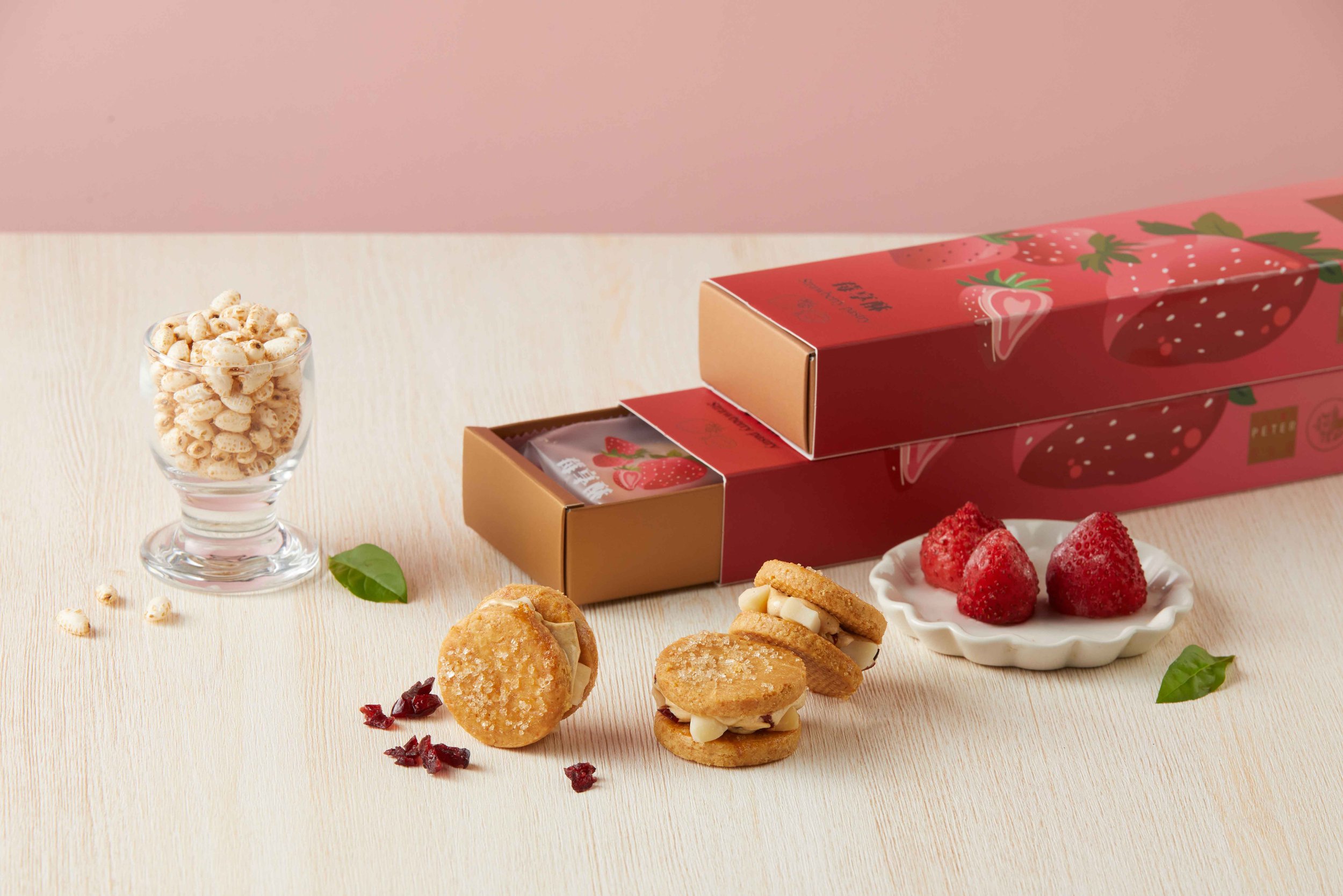 A glass bowl filled with puffed cereal, alongside strawberry-flavored snacks, cookies, strawberries, and dried cranberries on a light wooden surface with a pink background.