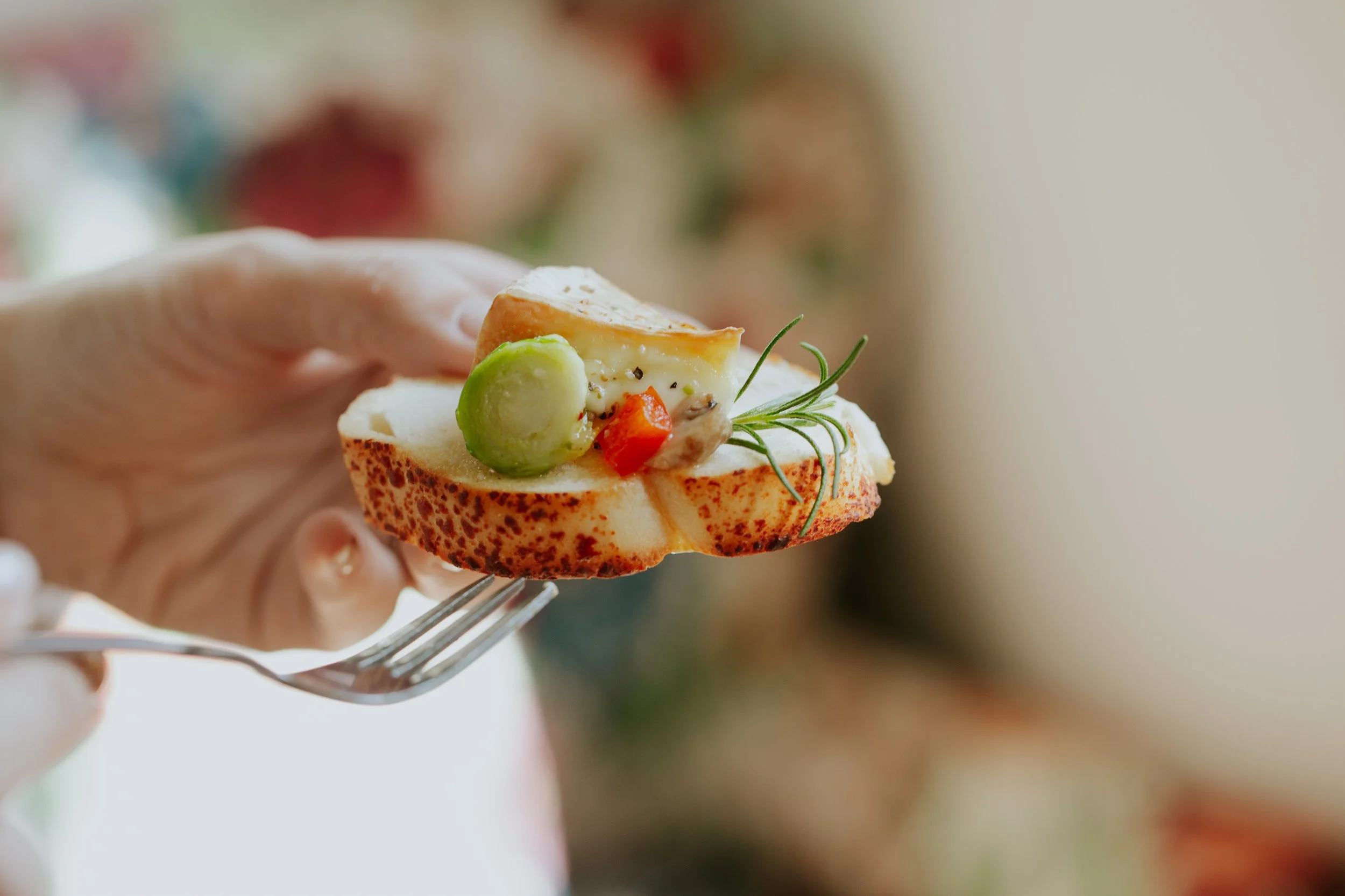 Close-up of a hand holding a fork with a slice of baguette topped with cheese, a green grape, a cherry tomato, and garnished with a sprig of rosemary.
