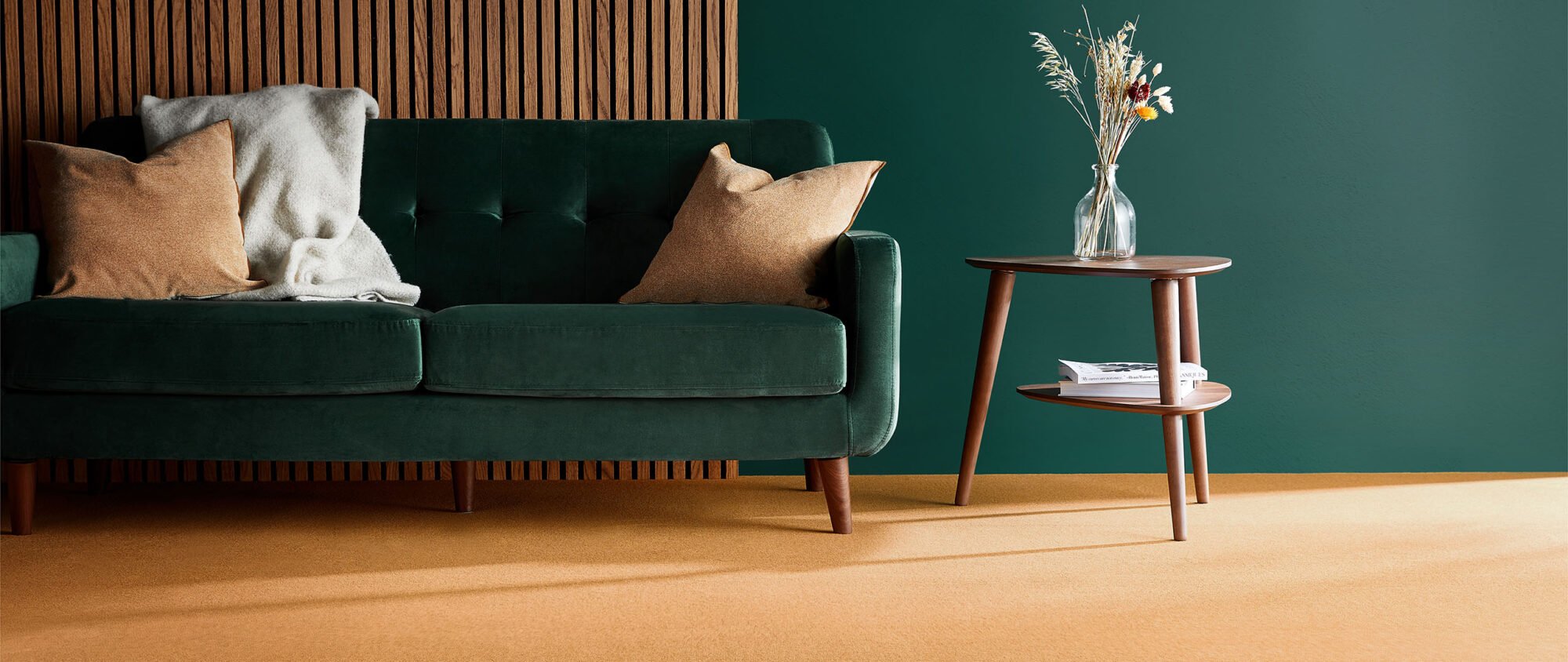 A modern minimalist room with a dark green painted wall and a section of the wall with thin oak slats running vertically. In front is a dark green sofa with oak legs and beside an oak coffee table with a vase of flowers on top.