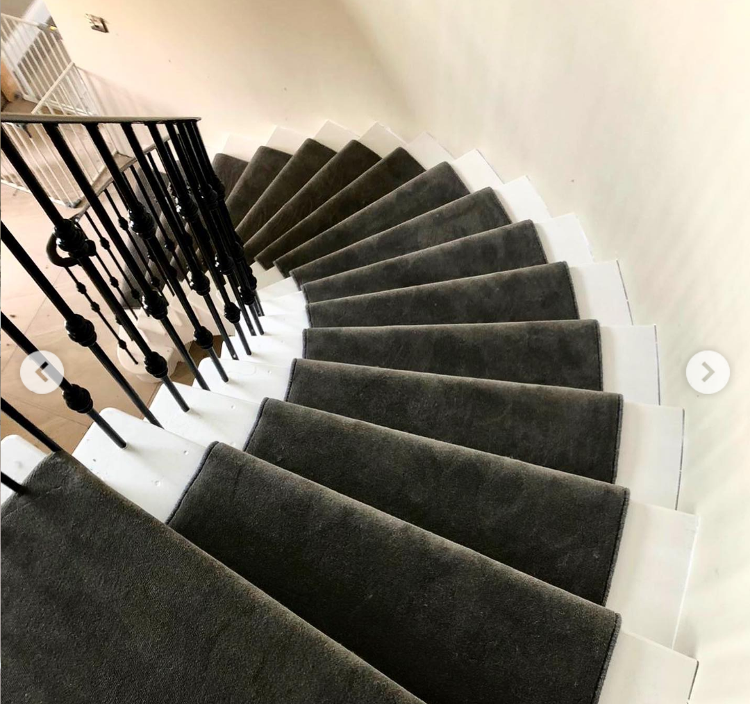 Looking down a curved white marble staircase with a dark velvet runner down the centre. On the left is a thin black metal handrail running downwards. Painted off white walls.