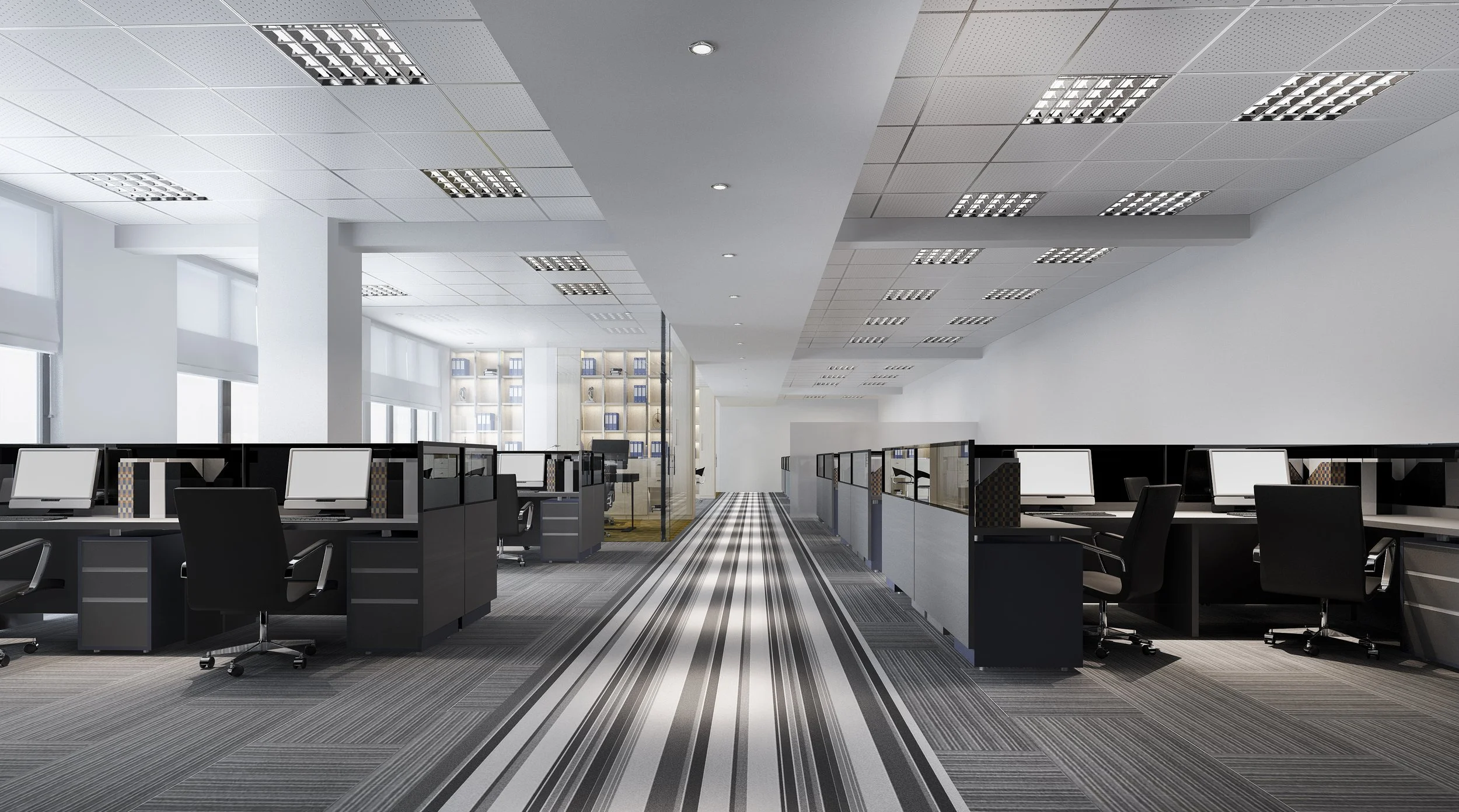 A modern office with rows of workstations, black desks, white monitors, and black office chairs. The ceiling has multiple square LED light panels, and the floor has a striped gray carpet. Large windows allow natural light, and there are bookshelves a