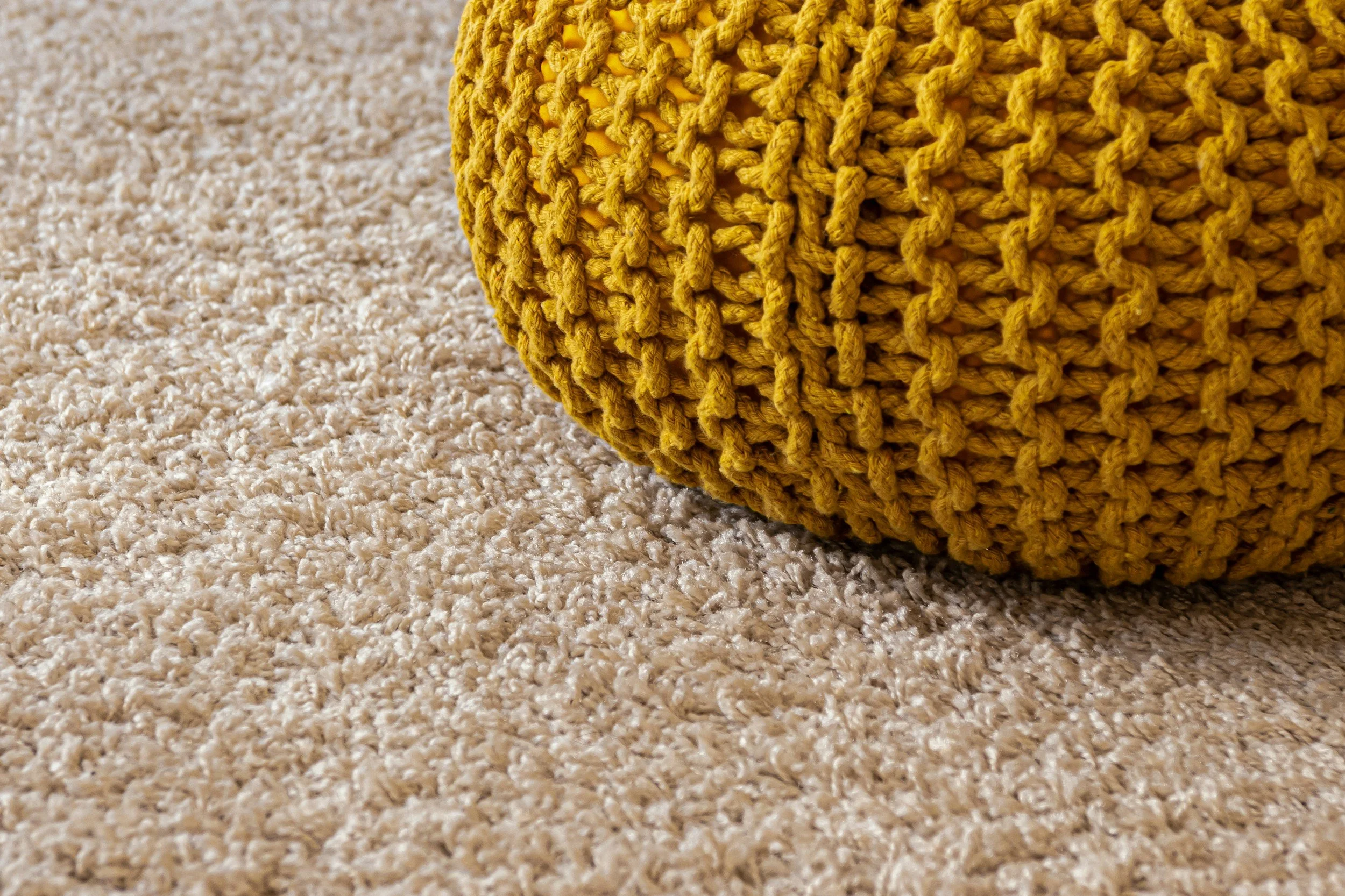 A close up image of a plush cream beige carpet and a mustard colour knitted round pouffe.