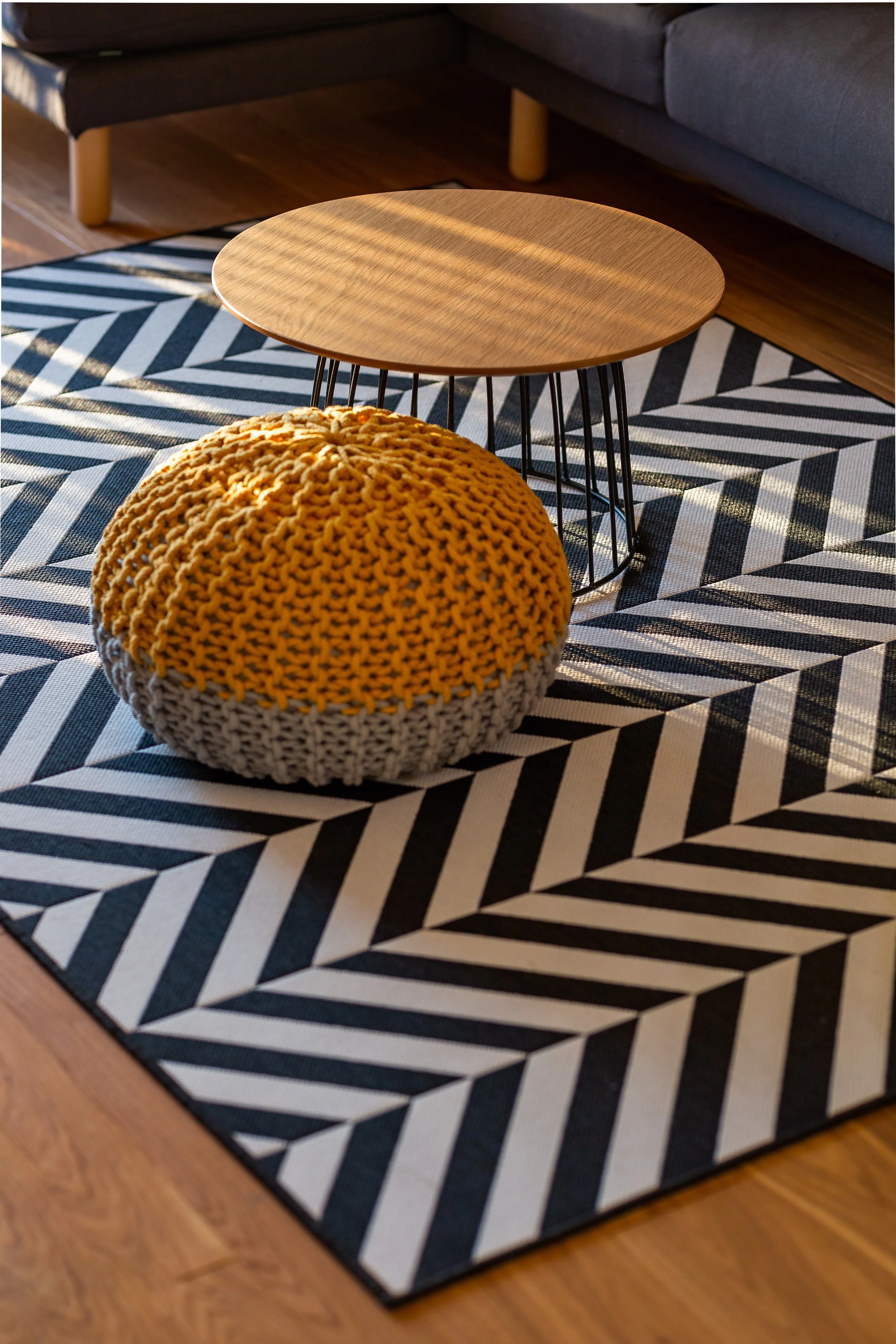 A black and white geometric patterned rug on a wooden floor in a living room. On the rug, there is a small round wooden table with black metal hairpin legs, and a large yellow knitted pouf in front of it. In the background, parts of a gray sofa and a dark coffee table are visible.