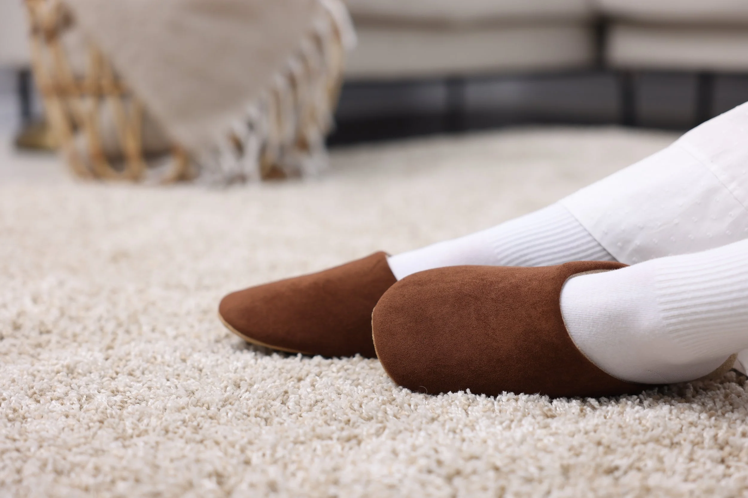 Person sitting on beige carpeted floor with legs extended, wearing brown shoes, white socks, khaki pants, and a white shirt, near a beige blanket with tassels and a metal bed frame.