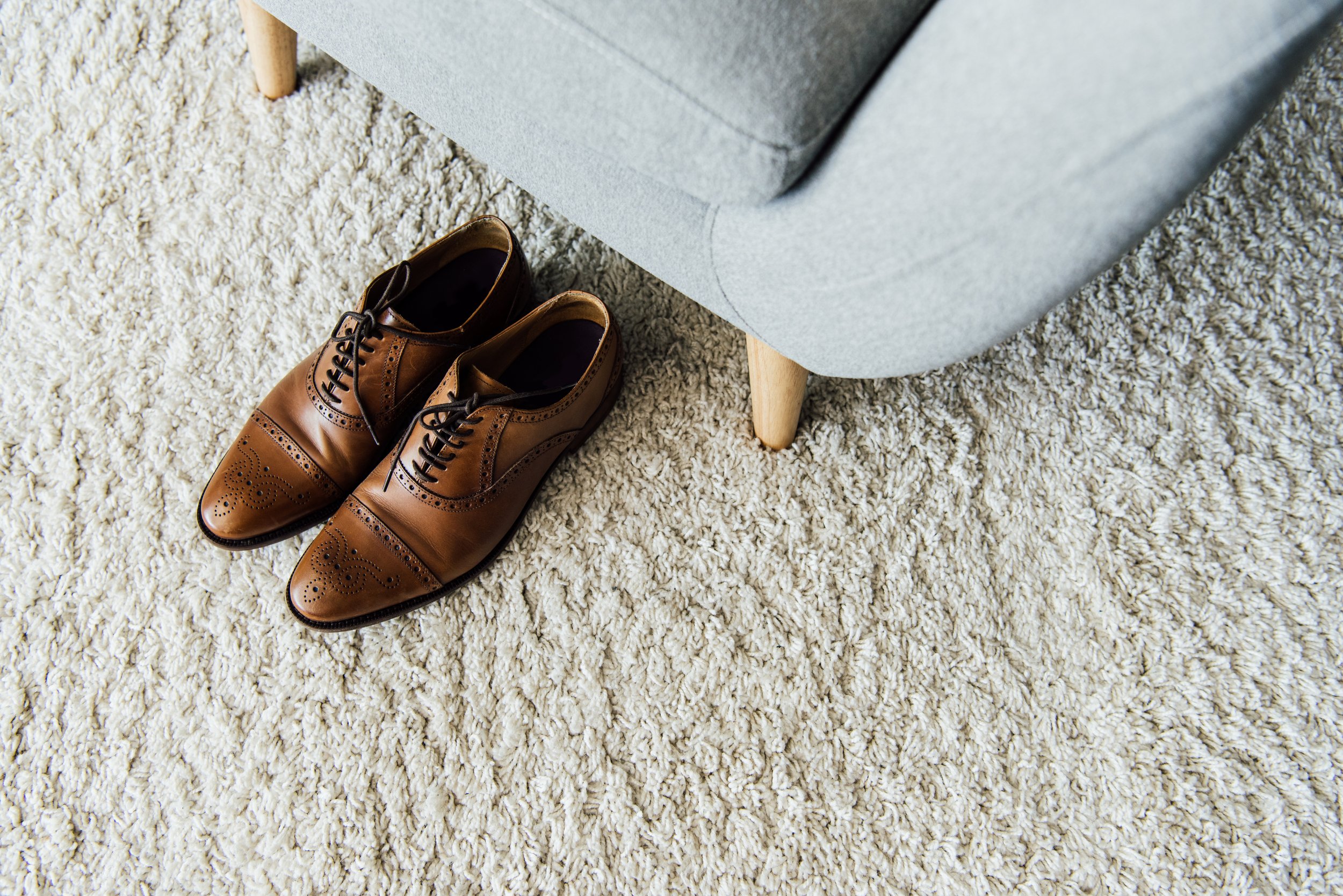 Brown leather dress shoes on a beige carpet next to a light gray armchair with wooden legs.
