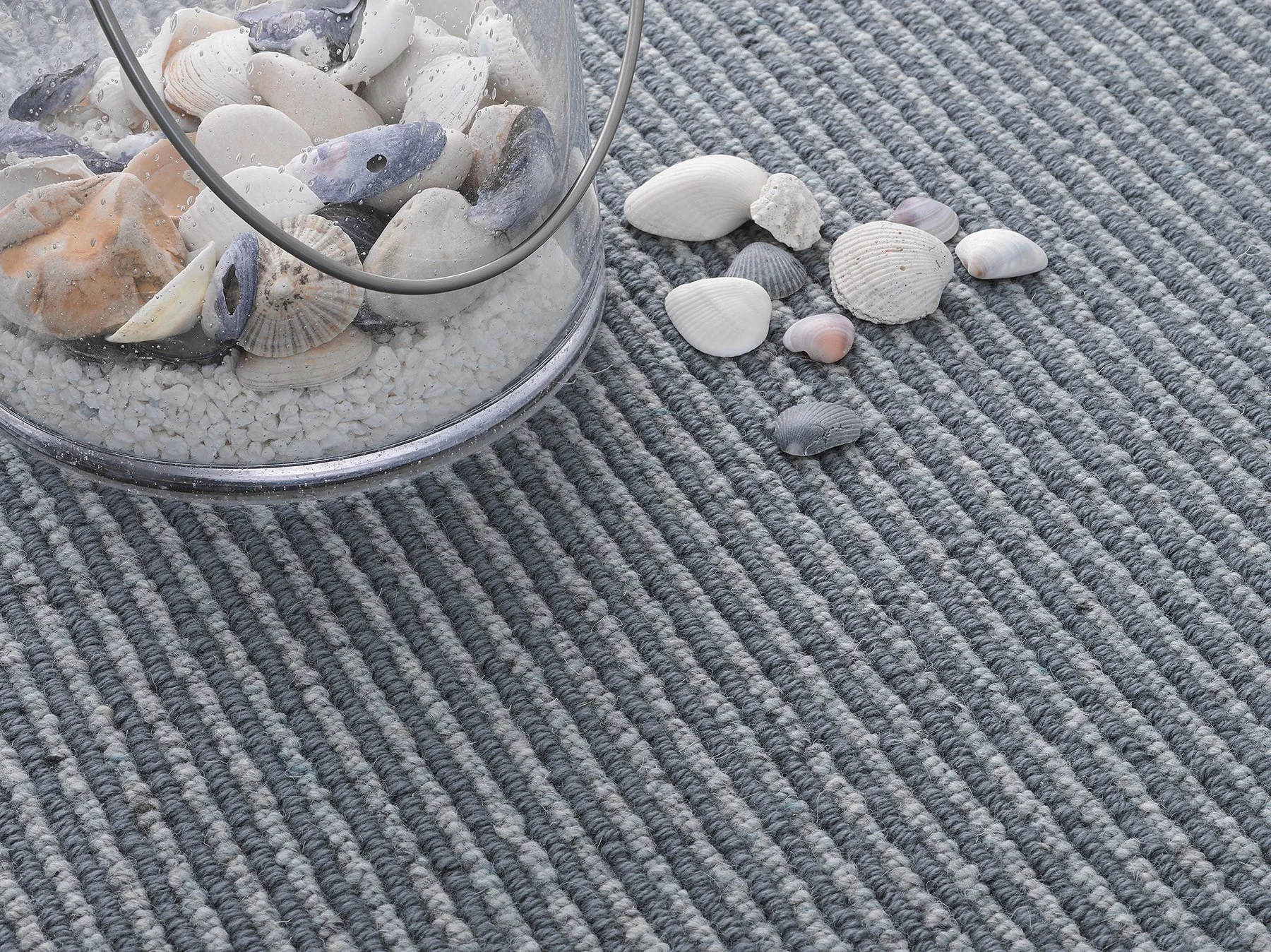 A blue grey loop pile carpet with line of alternating colour. On top is a glass vase with sand and shells within and some stones on the carpet giving a seaside vibe.