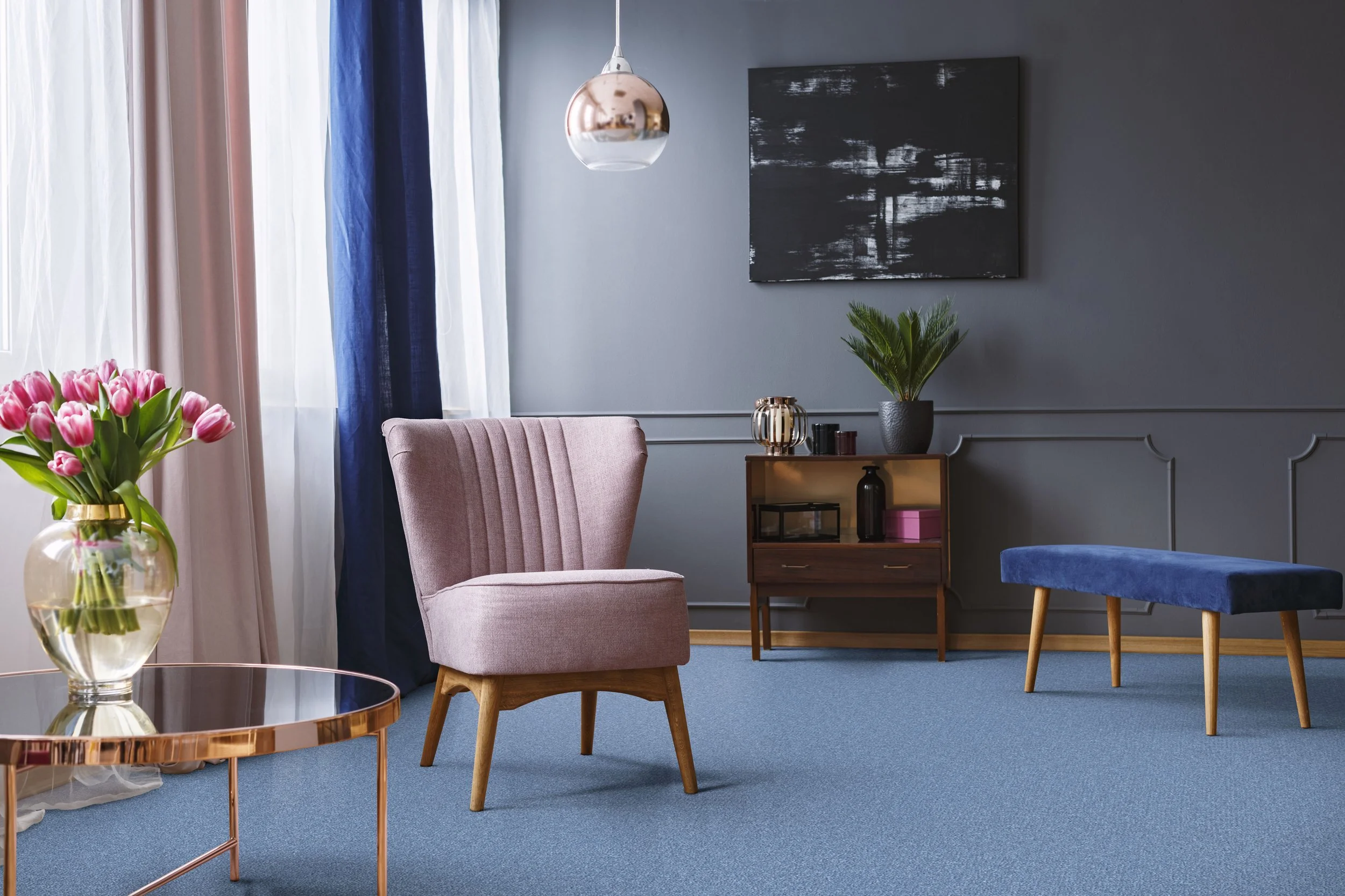 Stylish living area with muted blue carpet and walls. The room has a pink single armchair, a blue fabric footstool with wooden legs, a wooden cabinet agains the wall, and in the foreground a thin brass framed table on which there is a vase with pink 