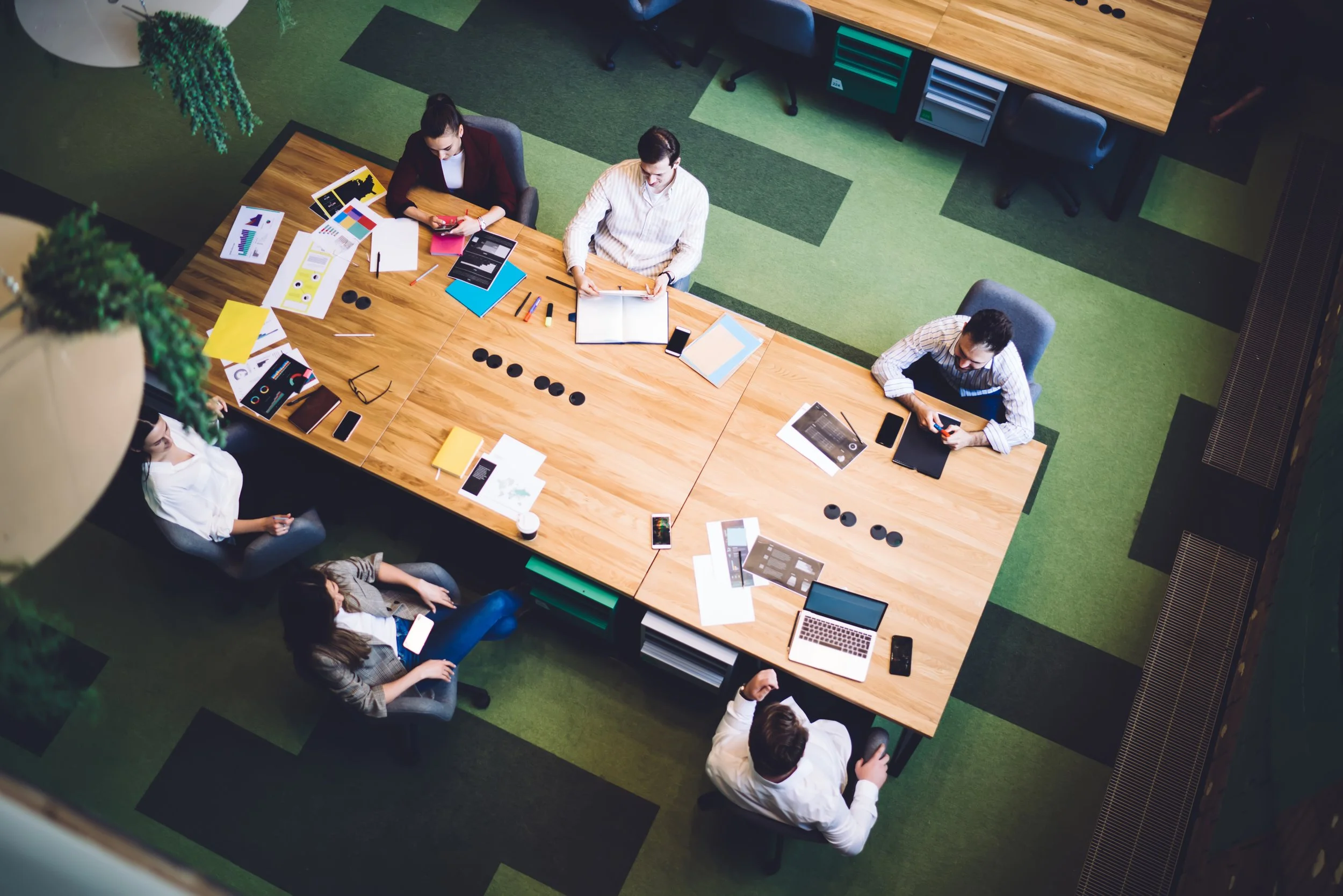 Overhead view of a meeting room with seven people sitting around a large wooden table, working on laptops, tablets, and smartphones, with documents and colored charts scattered on the table, and a green and black carpeted floor.