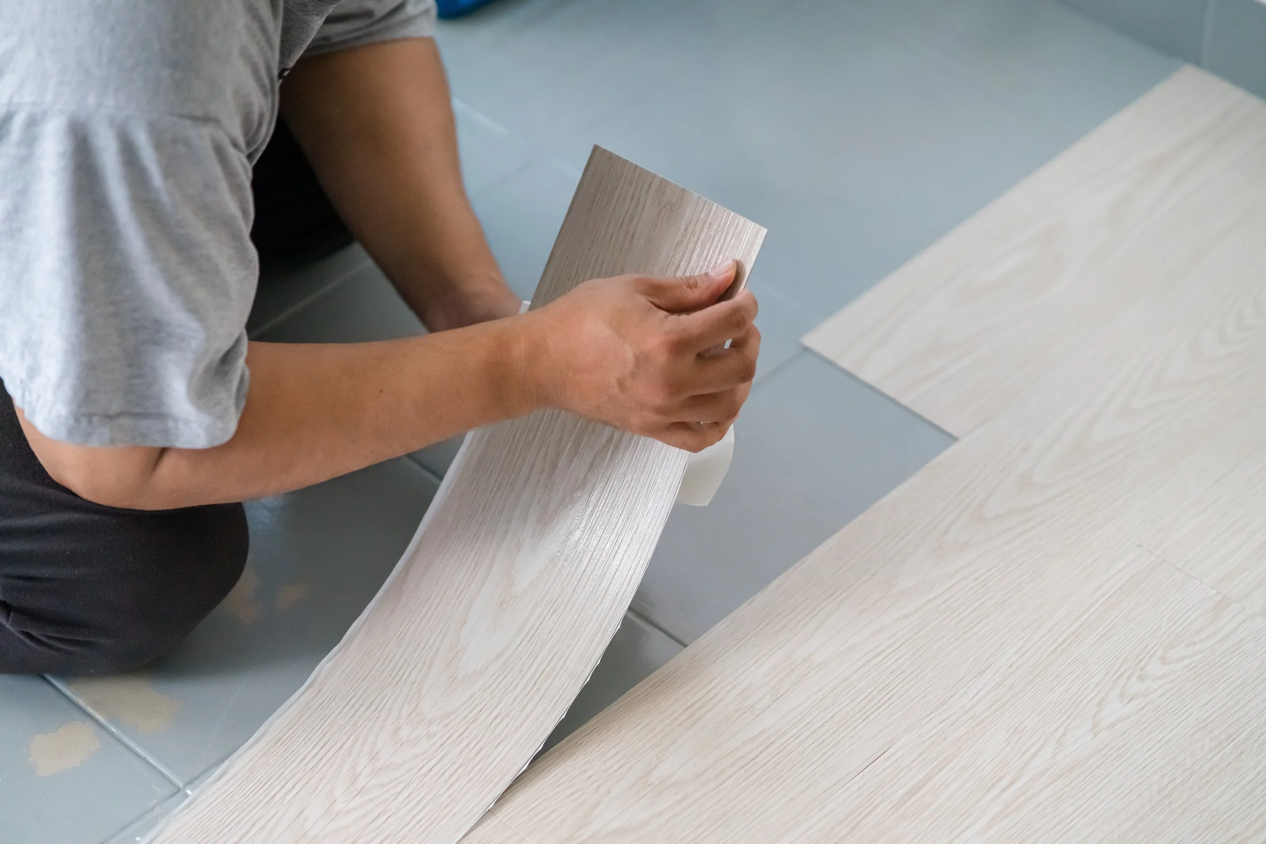 A person holding a piece of light-colored wood, standing next to a matching wooden surface.
