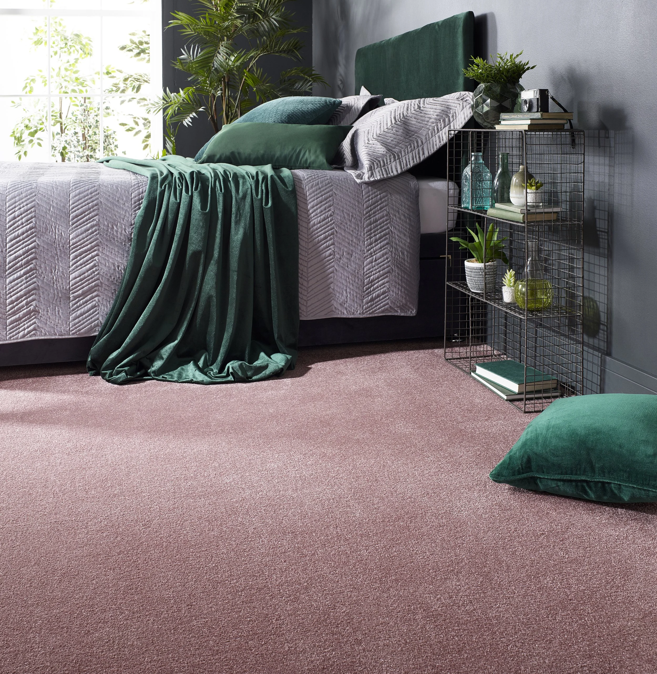 A stylish bedroom scene with dark green pillows and a throw on a grey bed. The wall behind is a light grey and in the foreground is a thin black framed shelving unit on which there are some books and small indoor plants with green foliage. TO the sid