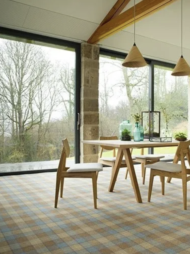 A dining area with a wooden table and four chairs with white cushions, large floor-to-ceiling windows showing trees outside, pendant lights, and decorative vases with plants on the table.