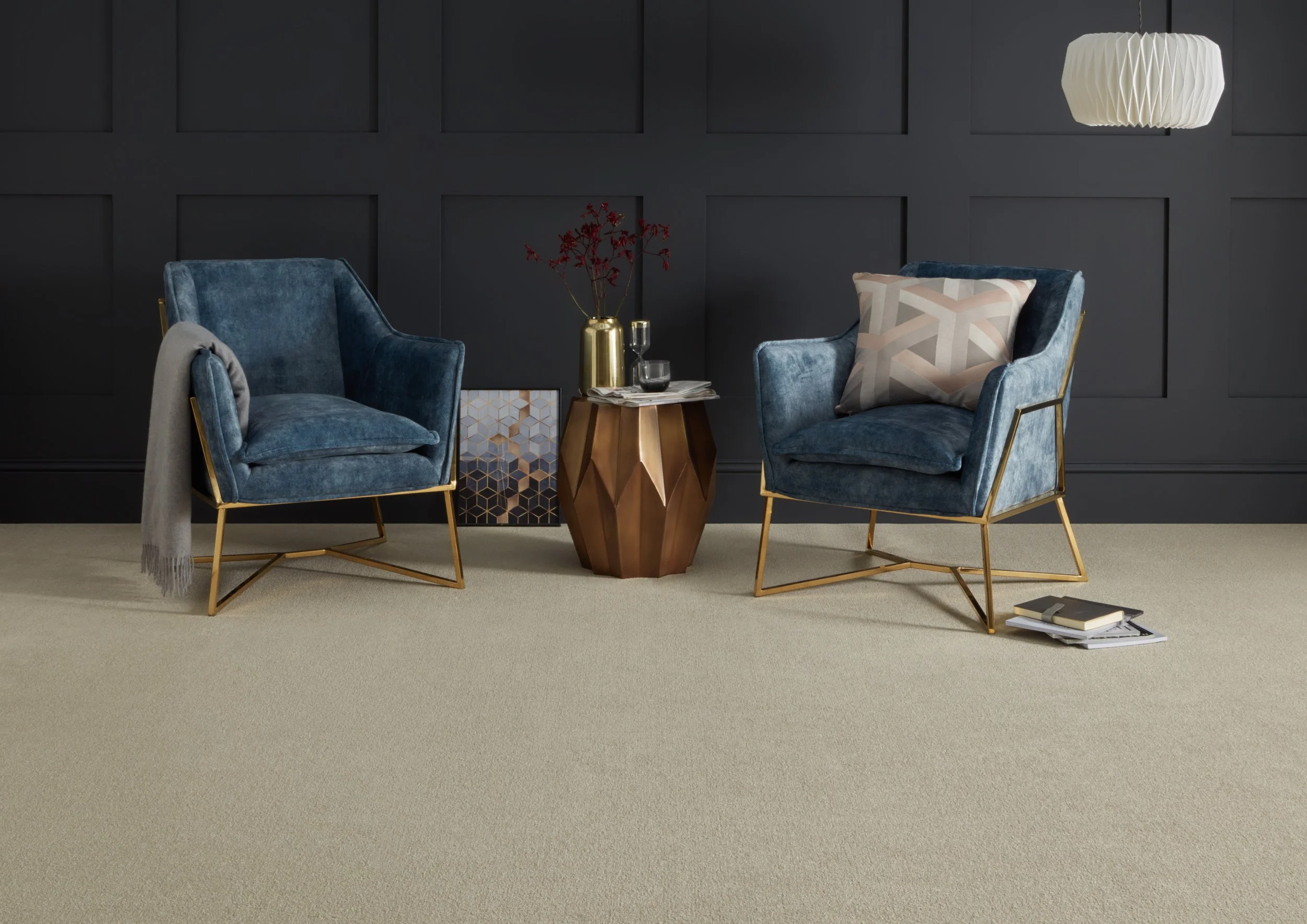 A chic living space with dark blue painted panelling behind and two single armchairs in blue suede with brass legs.. Between is a modern barrel shaped copper brass coffee table with a brass vase on top. Inside the vase are thin branches with dark red