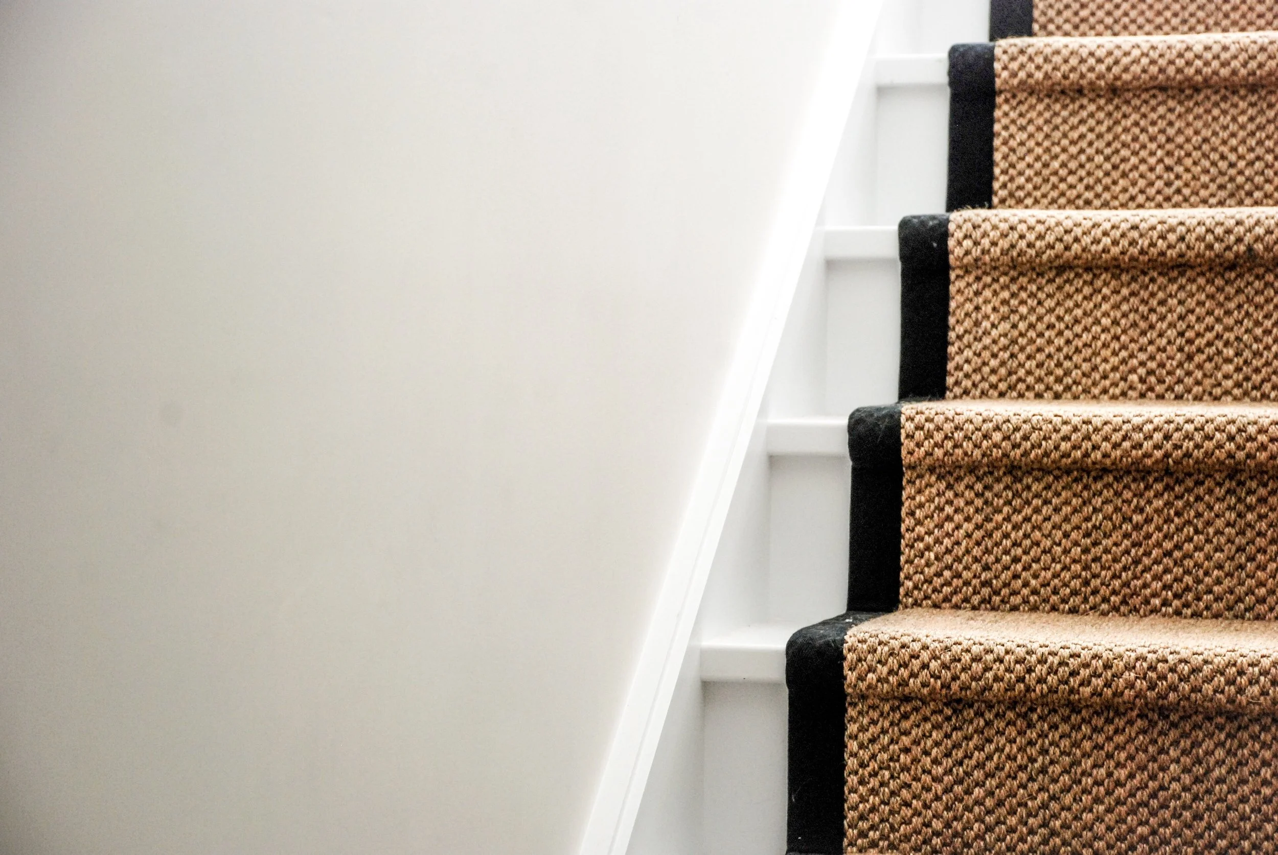 White painted staircase and off white painted walls with a natural fibre carpet runner and a thick black taped edging running down the staircase.