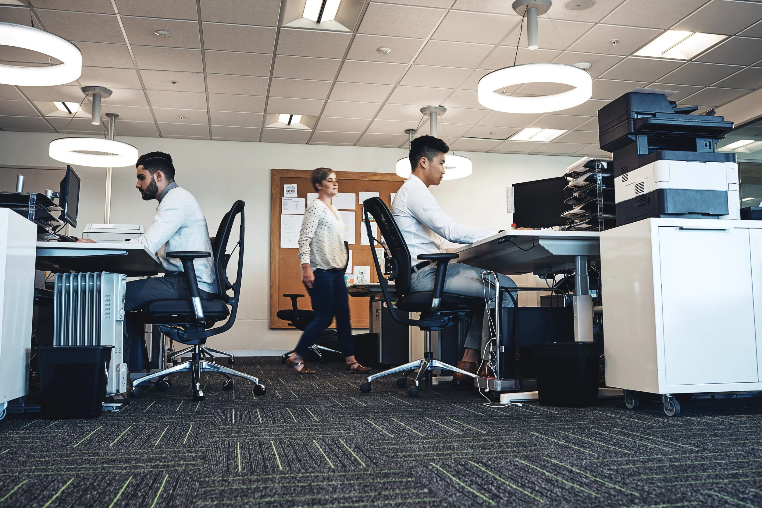 An office with three people working at desks, and one person walking by, under circular ceiling lights, with patterned carpeting.