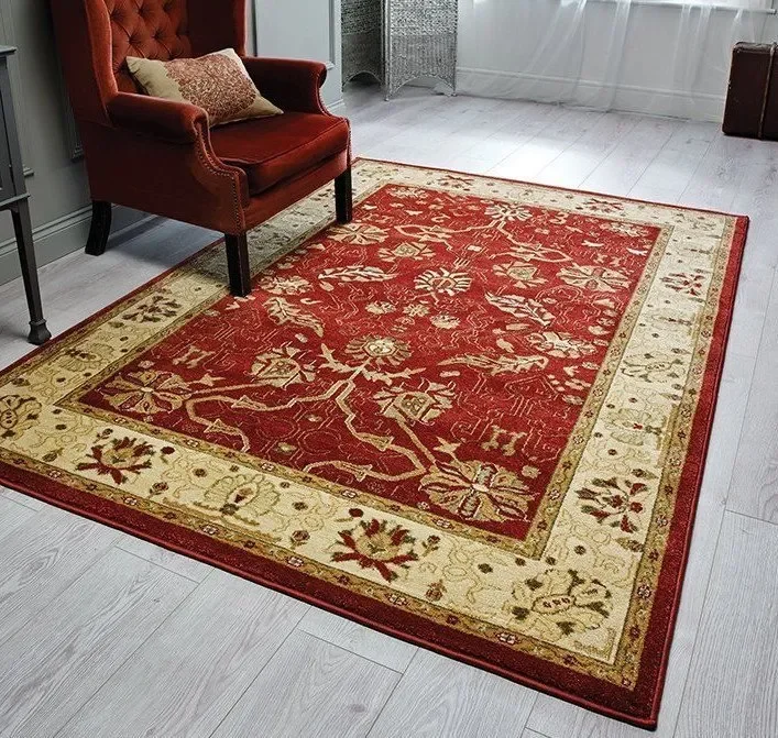 A red and beige area rug with floral and leaf patterns in a living room with light wood flooring, a red armchair with a decorative pillow, and a white curtain in the background.