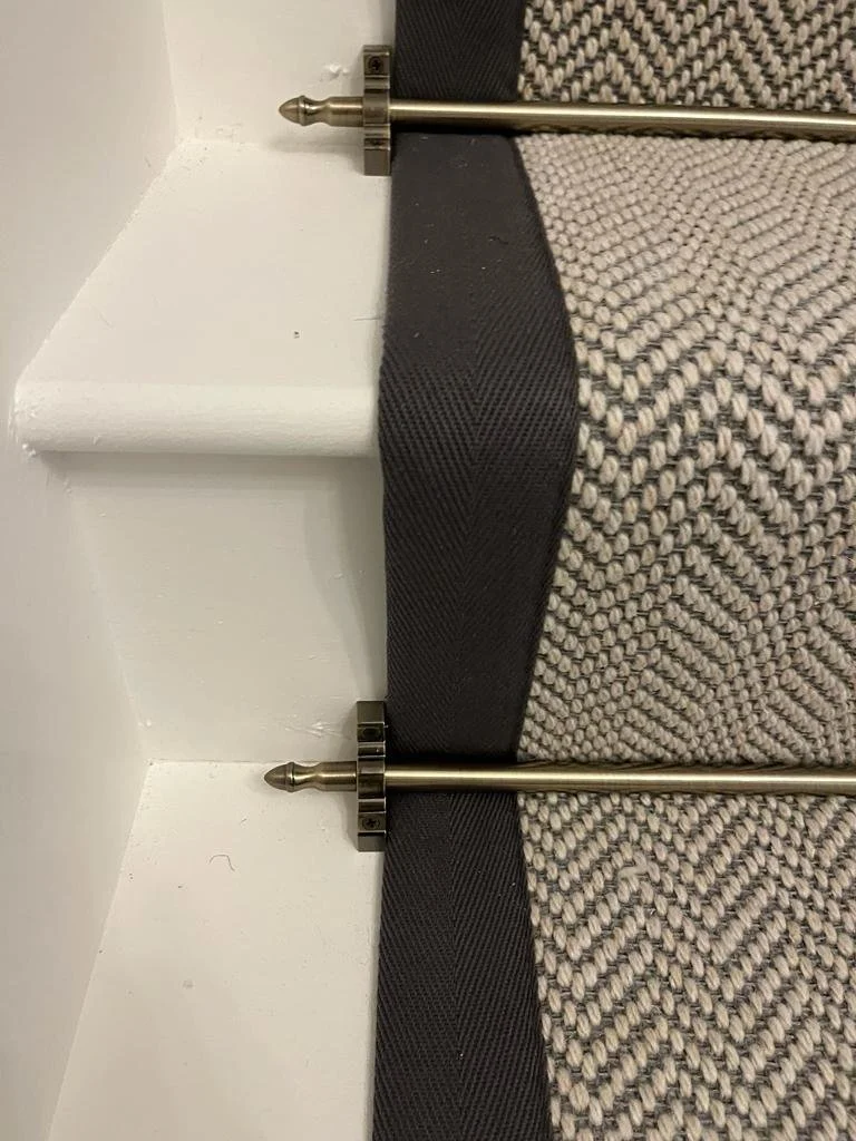 Close up of a herringbone design stair runner with a dark grey taped edging and a brass stair rod with white painted stairs.