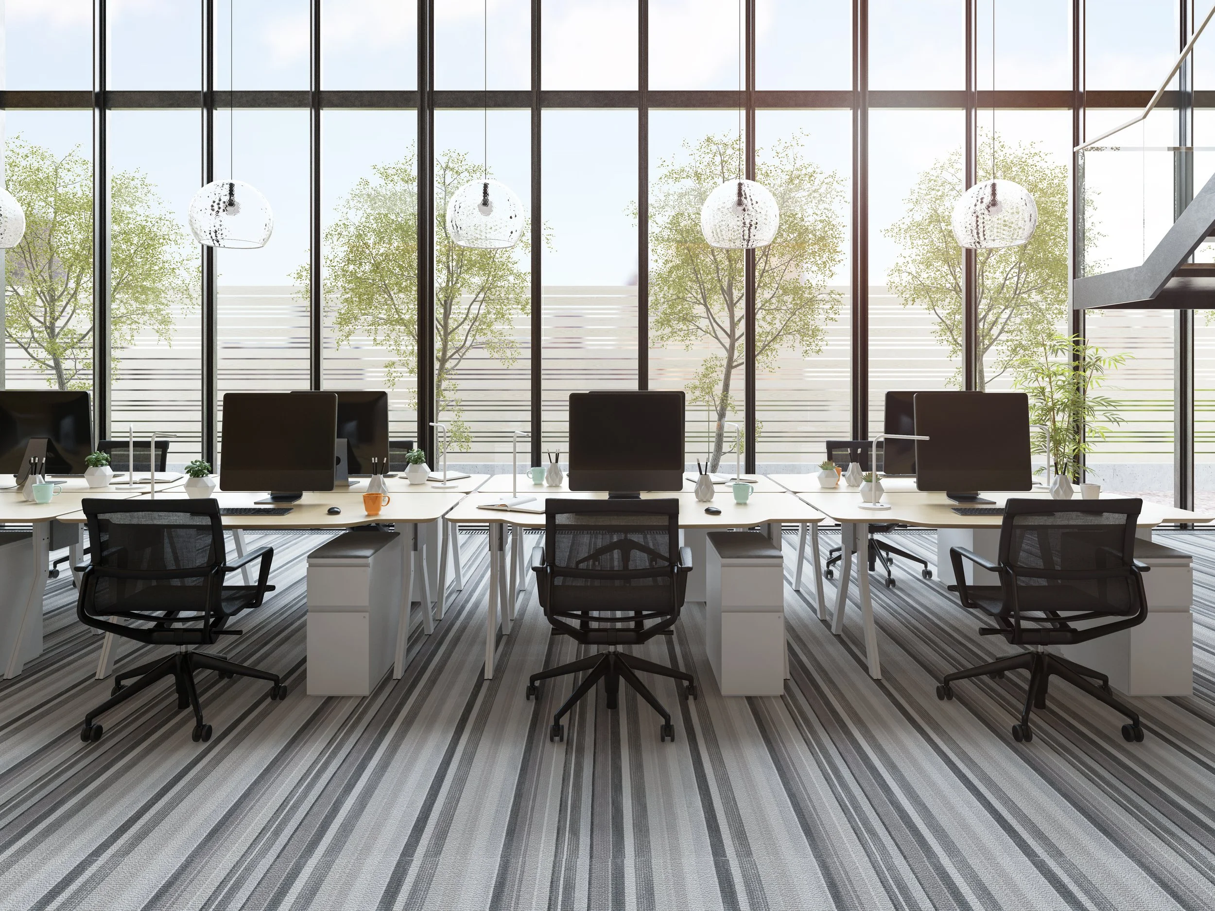 Modern office interior with large floor-to-ceiling windows, multiple workstations with computers, black office chairs, hanging pendant lights, and a striped carpet. Outside, trees are visible through the windows.