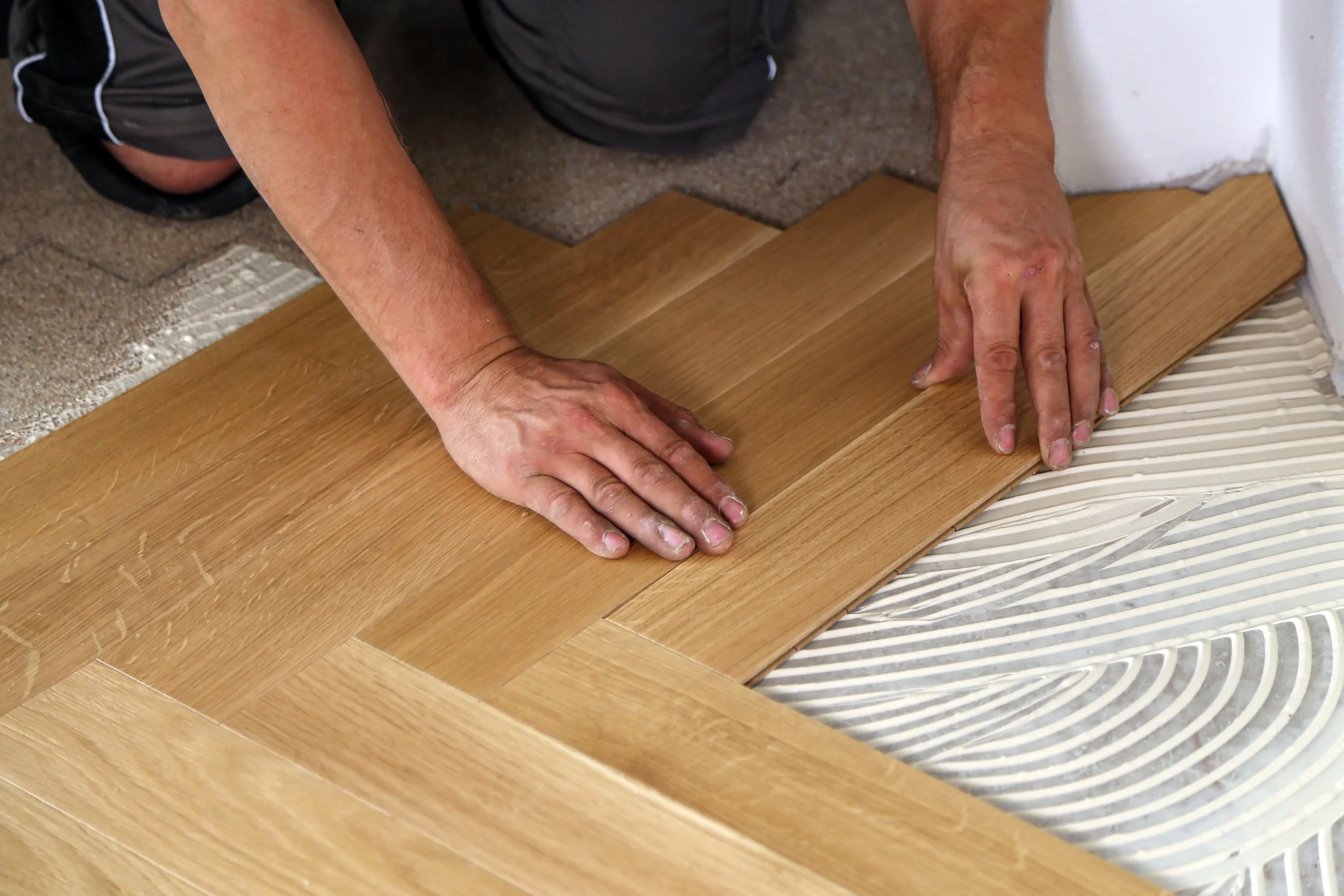 Person installing wooden flooring, kneeling on concrete subfloor, pressing hardwood planks into place.