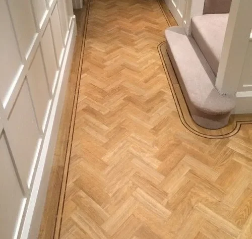 View of a hallway with light wood parquet flooring and a carpeted staircase at the top right corner.