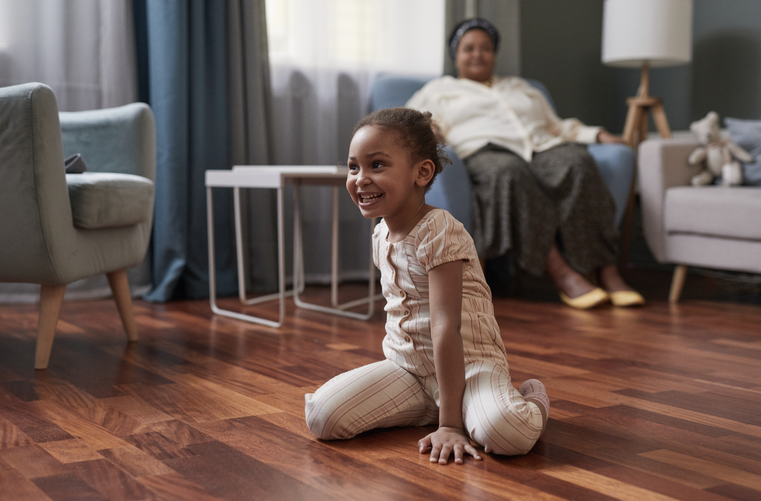 A young girl with braided hair, wearing beige and white striped pajamas, kneeling on a wooden floor, smiling and looking to her left, in a living room with a woman sitting on a couch in the background.