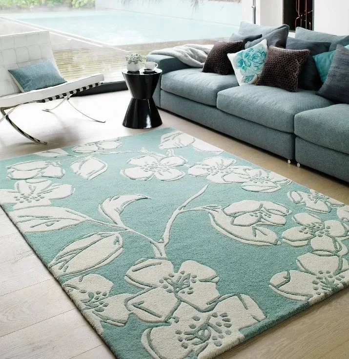Living room with a light blue floral patterned rug, a gray sofa with assorted cushions, a black side table with a plant and mug, and a white modern lounge chair near a large window.
