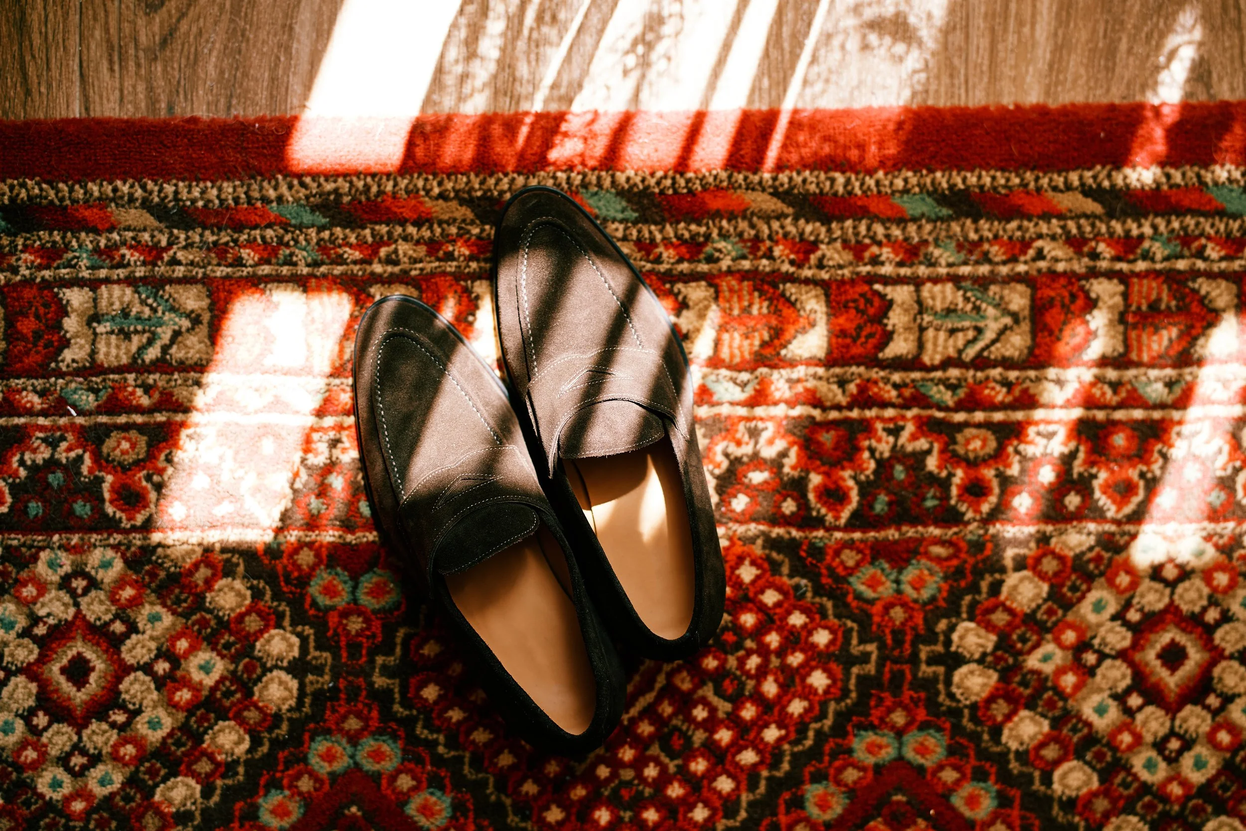 A pair of black leather shoes on an oriental patterned rug, with sunlight streaming through window blinds creating striped shadows.
