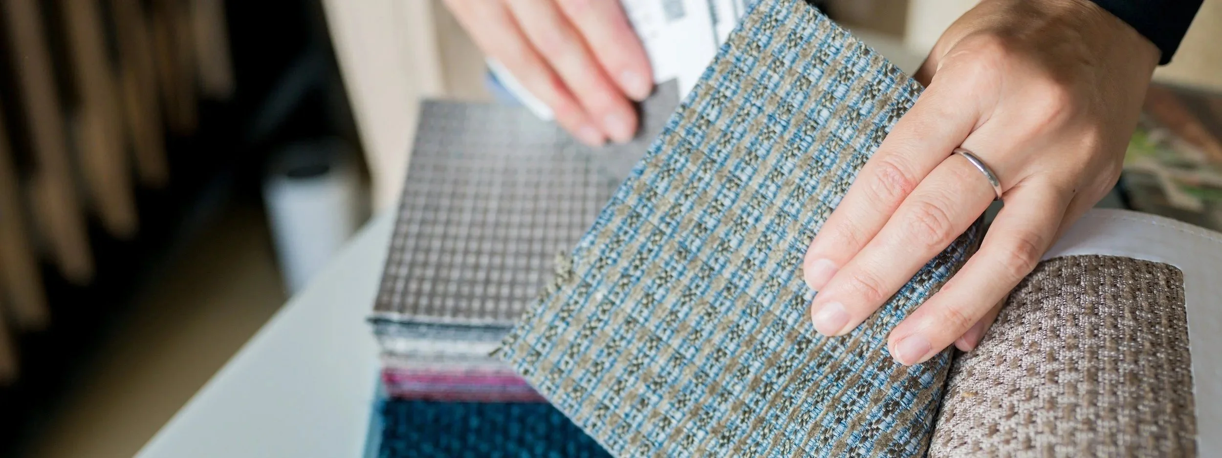 Close-up of a person's hand with a ring, holding and flipping through fabric samples of different textures and colors, including beige, gray, and brown.