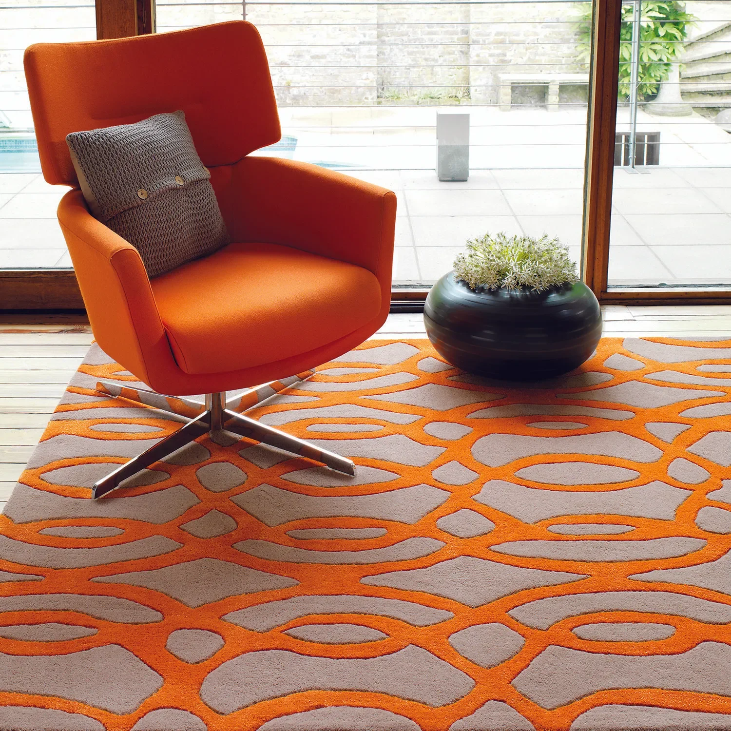 Modern orange armchair with a gray knitted pillow on a colorful orange and beige patterned rug, next to a glass sliding door leading to an outdoor patio with plants and a stone wall.