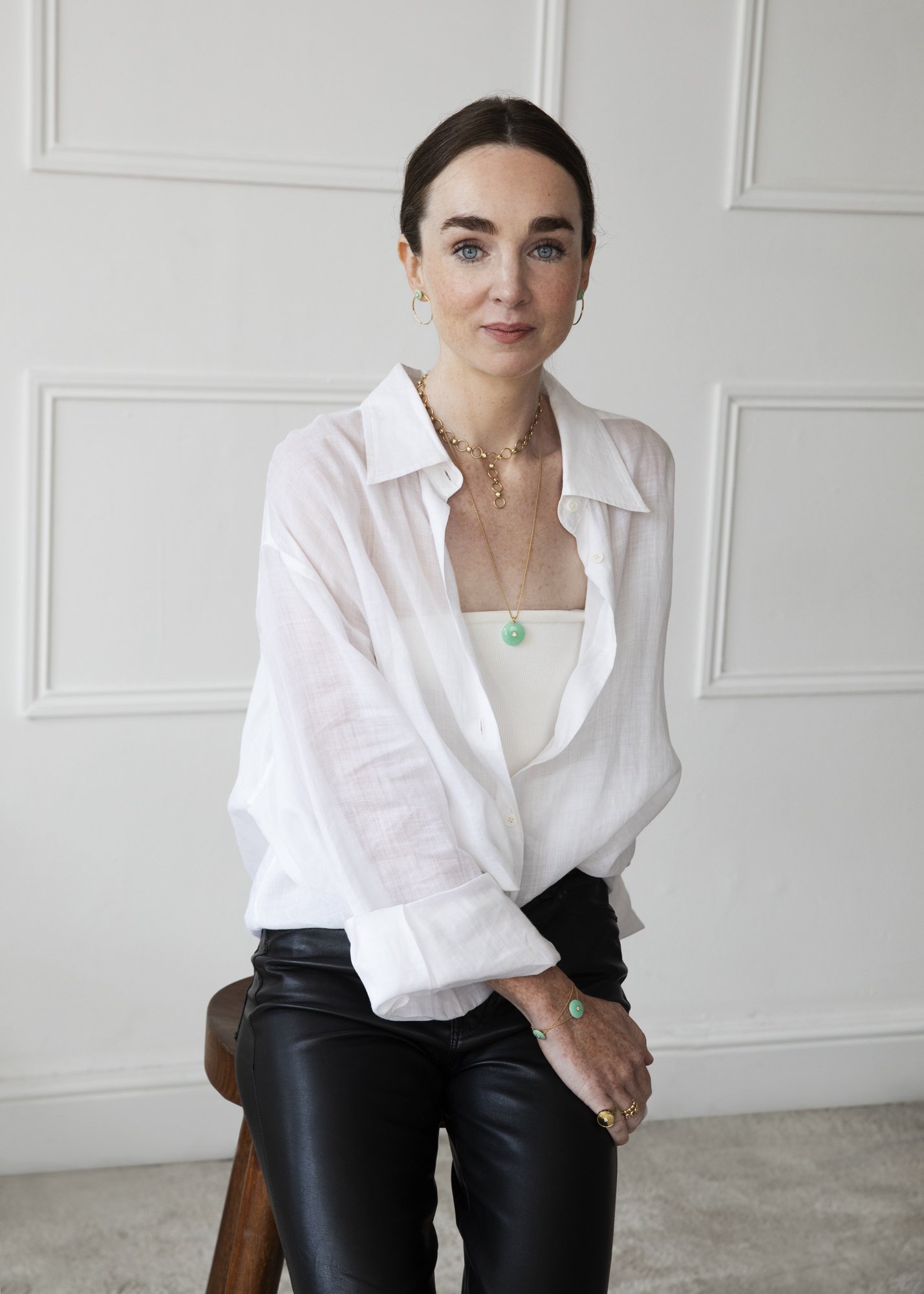 A woman with dark brown hair and blue eyes, wearing a white button-down shirt, black leather pants, and jewelry including gold rings, earrings, and layered necklaces with a green pendant, sitting on a wooden stool in front of a white wall with decorative molding.