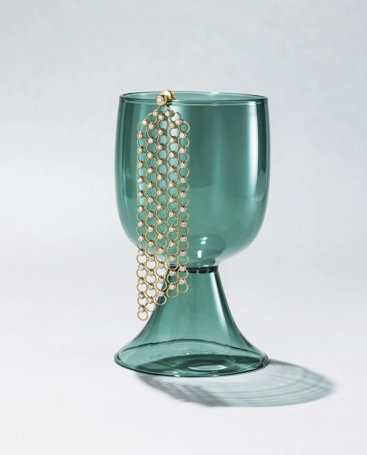 A green glass goblet with a gold and diamond bracelet draped over its edge, casting a shadow on a white background.
