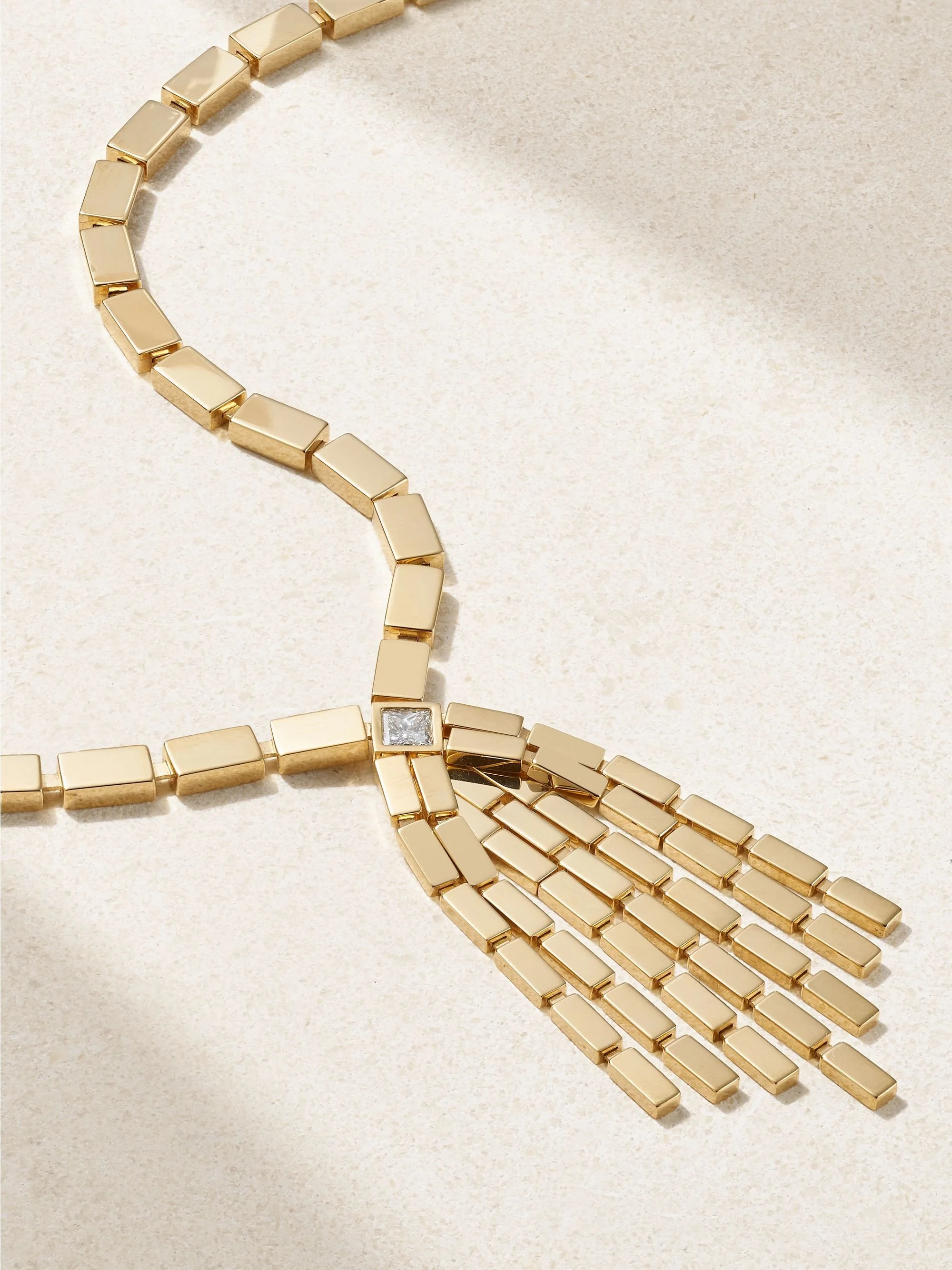 Close-up of a gold bracelet and layered necklace with rectangular links, including one with a square diamond accent, on a beige surface.