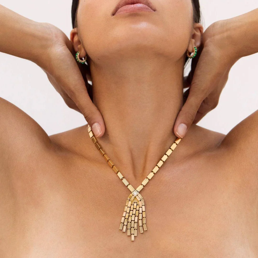 A woman wearing a gold necklace and earrings, with her hands resting on her neck, showing her bare shoulders and upper chest.