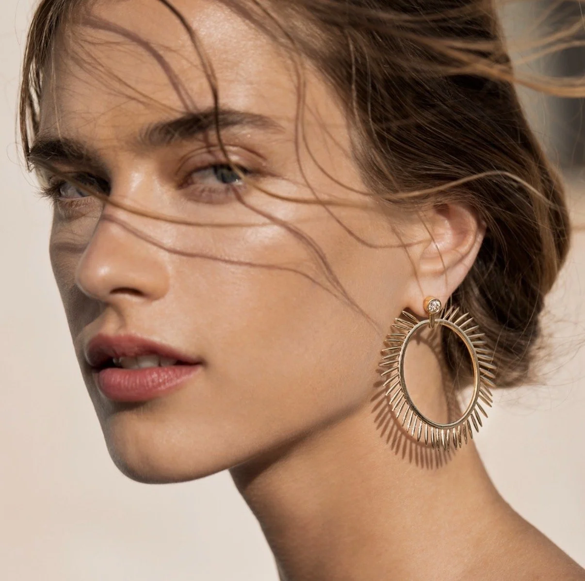 Close-up of a woman's face with wind-blown hair, wearing a large gold hoop earring with a sunburst design and a small diamond earring.