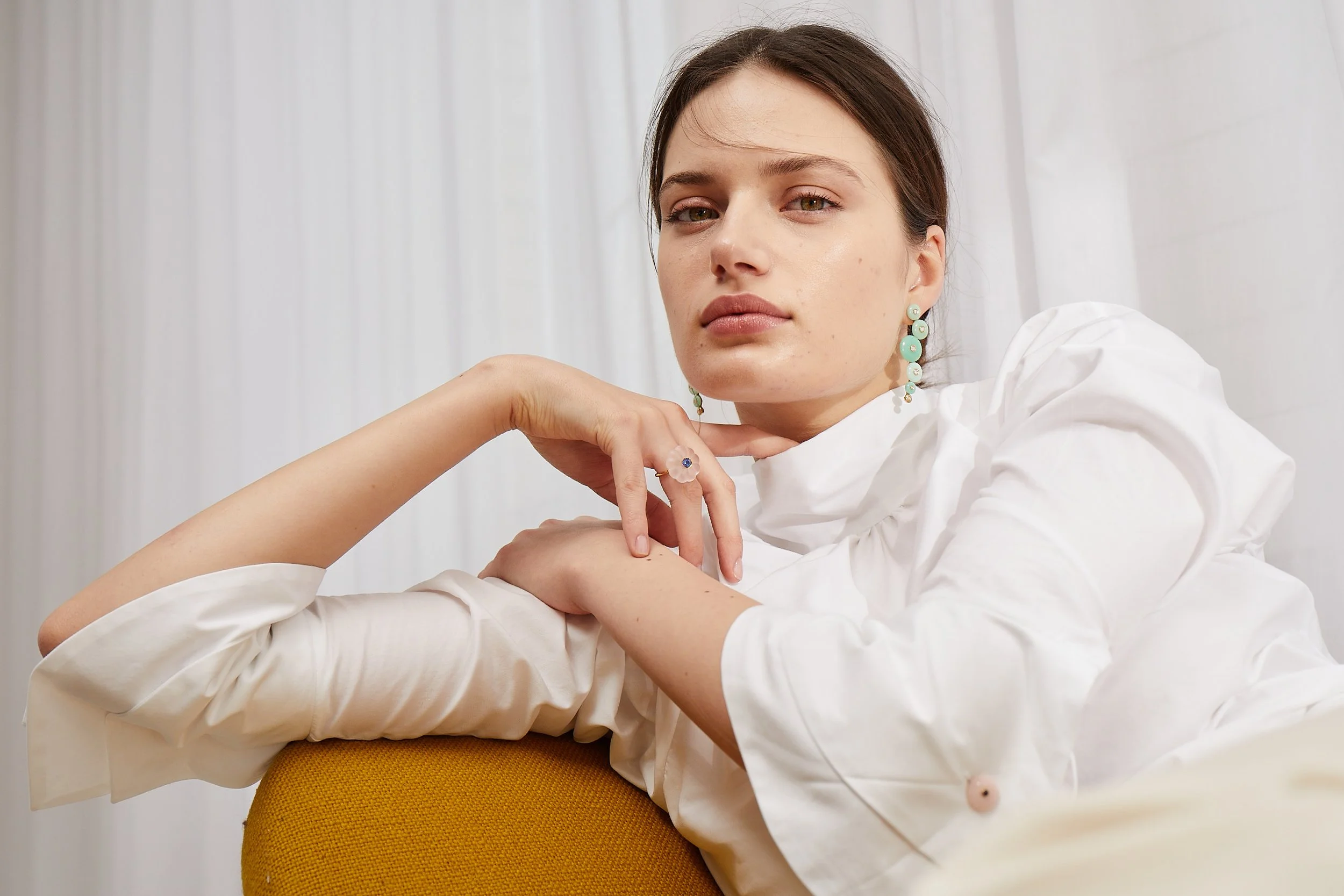 A woman with brown hair and light skin, wearing turquoise earrings and a white blouse, resting her chin on her hand, sitting in front of a white curtain.