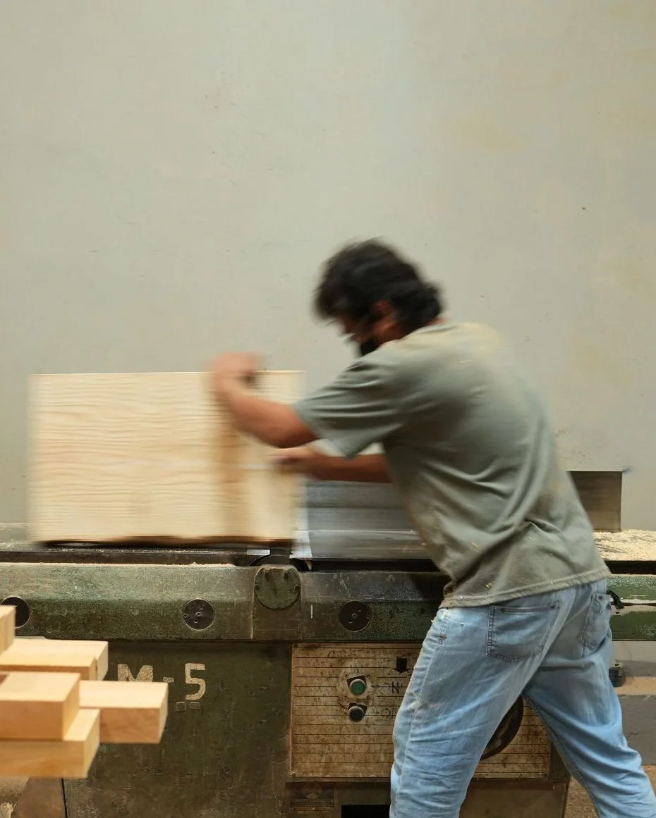 A man with dark hair and glasses working with a large woodworking saw, cutting a piece of wood in a workshop.