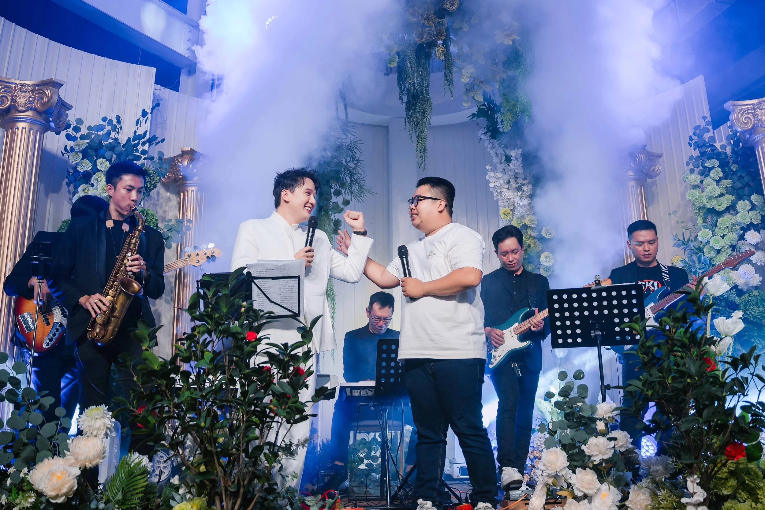 A group of musicians performing on stage, with two men in white shirts dancing and holding microphones, surrounded by flowers and blue lighting, with smoke effects.