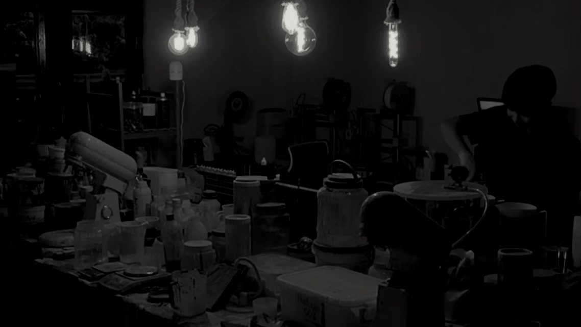 A dark room filled with various laboratory equipment and containers, illuminated by a few hanging light bulbs, with a person working at the right side.