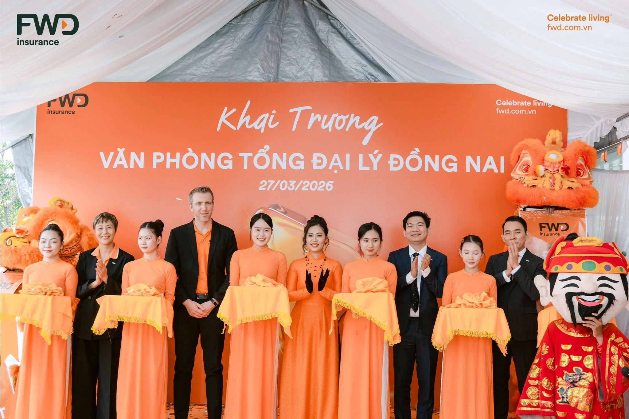 Group of people celebrating the grand opening event with a large orange banner behind them, featuring the date 27/03/2026 and the Vietnamese words 'Khai Trương Văn Phòng TỔNG ĐÀI ĐỒNG NAI', alongside lion dance costumes and performers.