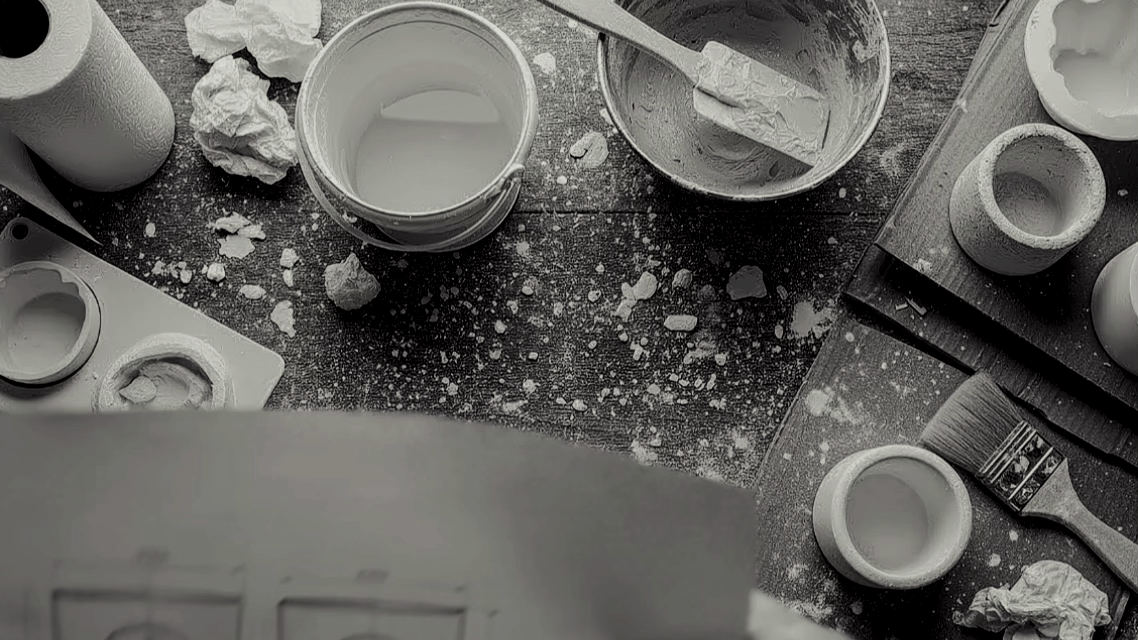 A messy baking workspace with flour scattered on a dark surface, eggshells, bowls, a paintbrush, and baking tools.