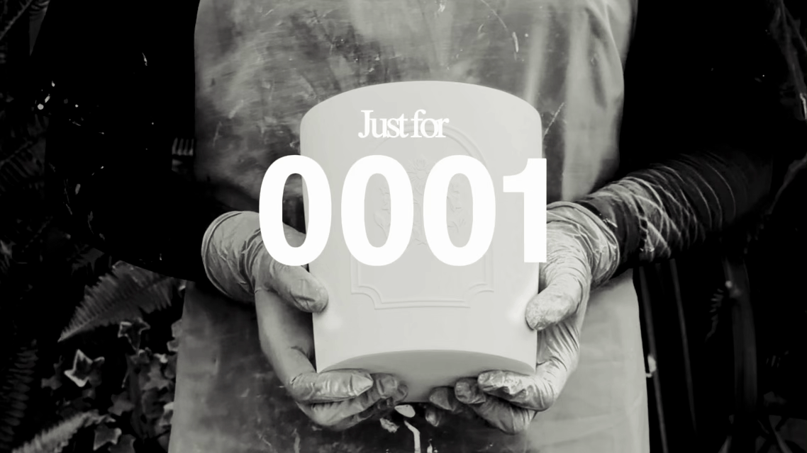 A person wearing gloves holding a white rectangular object with 'Just for 0001' written on it, in black and white.