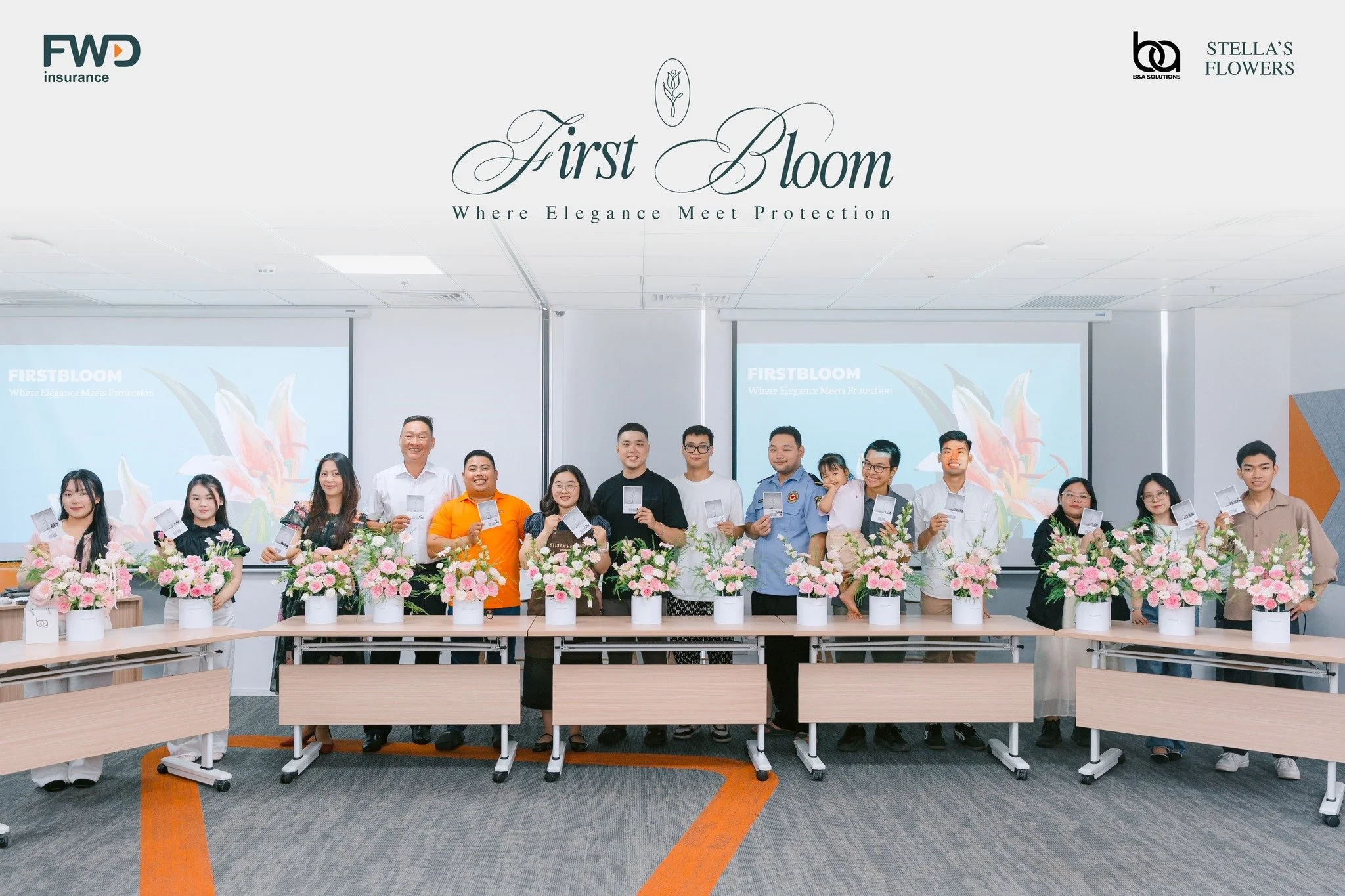 Group of people standing behind a row of pink and white flower arrangements on desks, in a conference or presentation room with screens and a large sign that reads 'First Bloom, Where Elegance Meets Protection'.