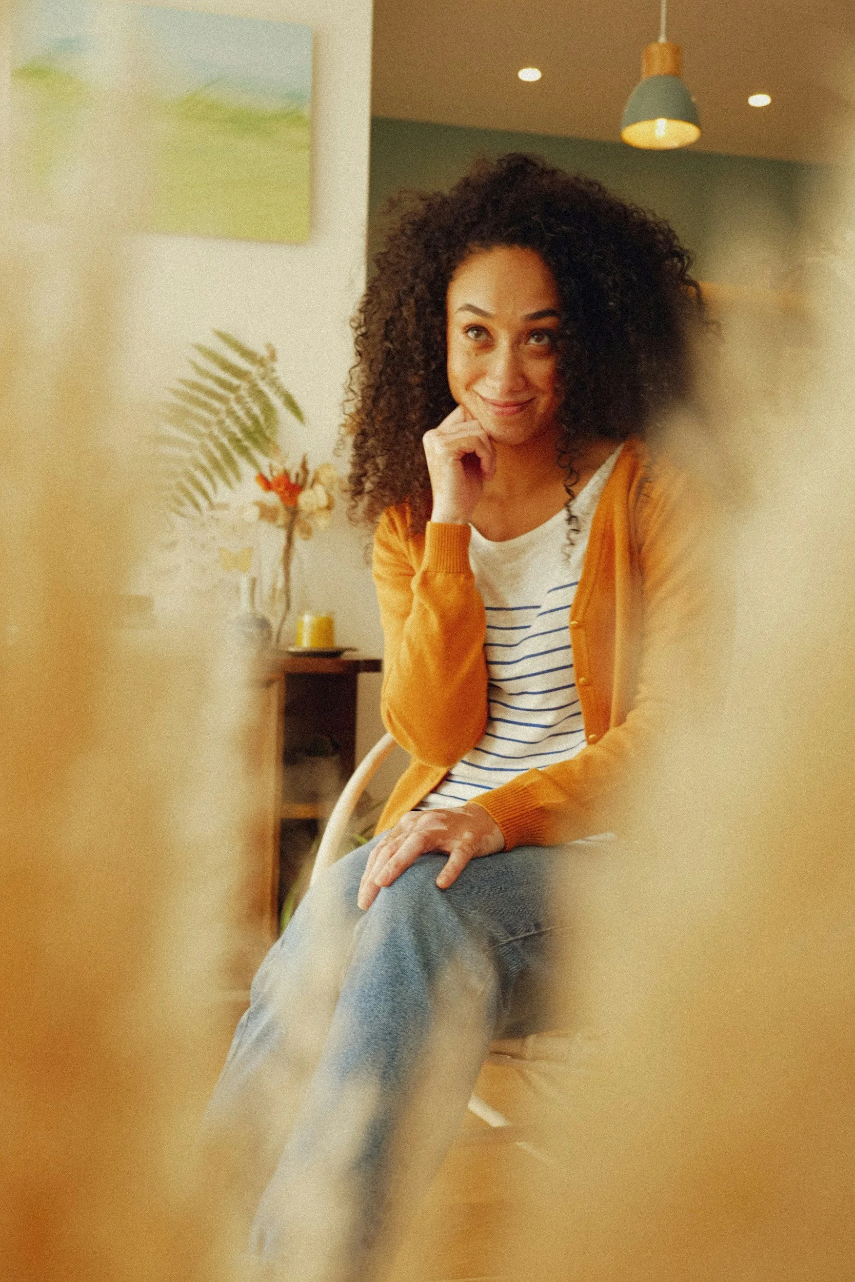 A woman with curly hair sitting indoors on a chair, wearing a striped shirt and a mustard yellow cardigan, smiling and resting her chin on her hand.