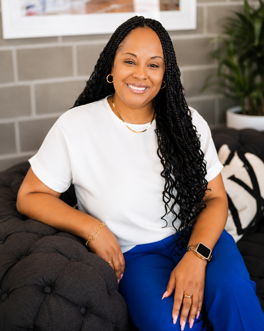 A smiling woman with long black braids, wearing a white shirt and blue pants, sitting on a black tufted sofa in a modern room with a gray brick wall and a potted plant in the background.