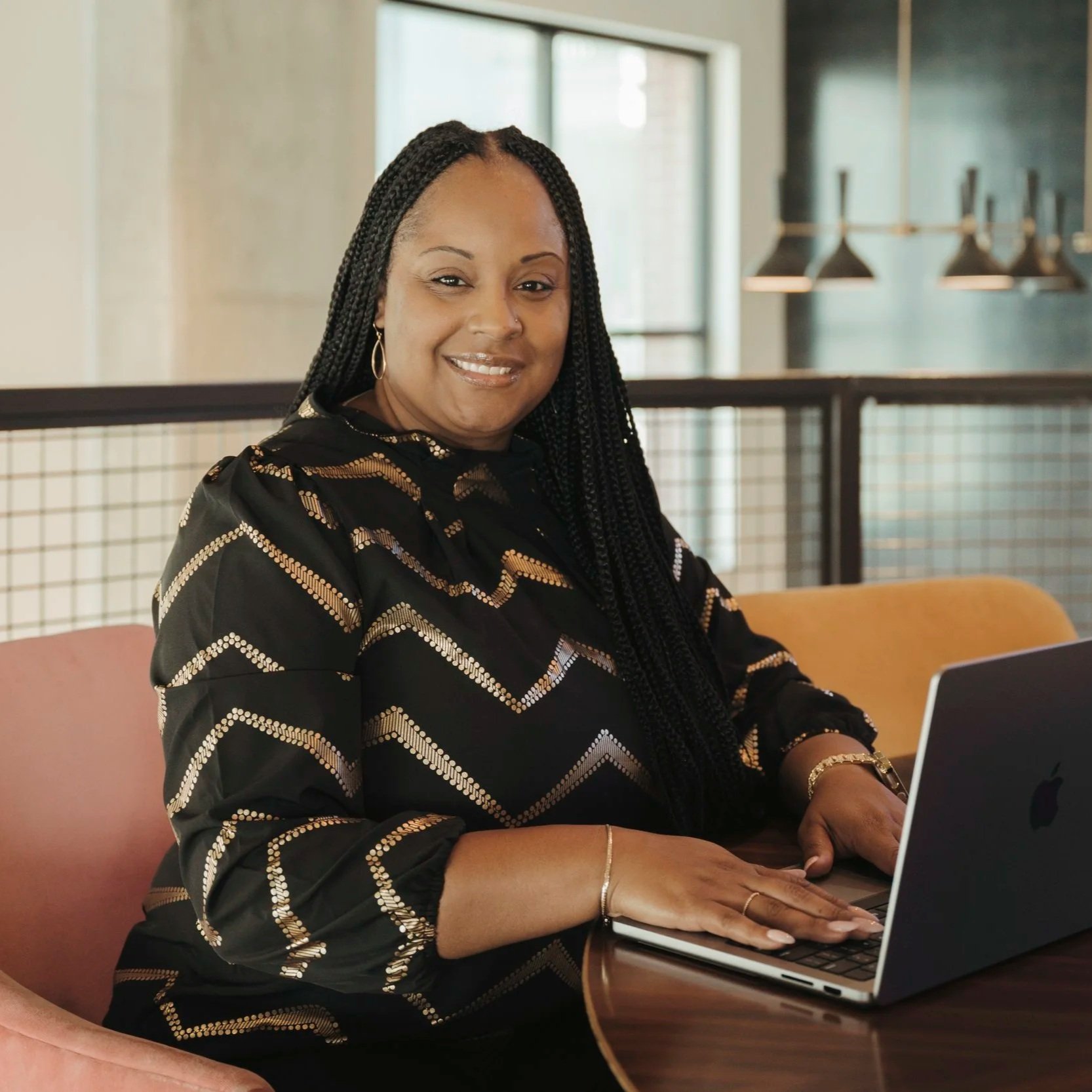 A woman with braided hair wearing a black and gold patterned top, sitting at a wooden table with a laptop in front of her. She is smiling and appears to be working or studying in a modern indoor space with large windows and hanging lights.