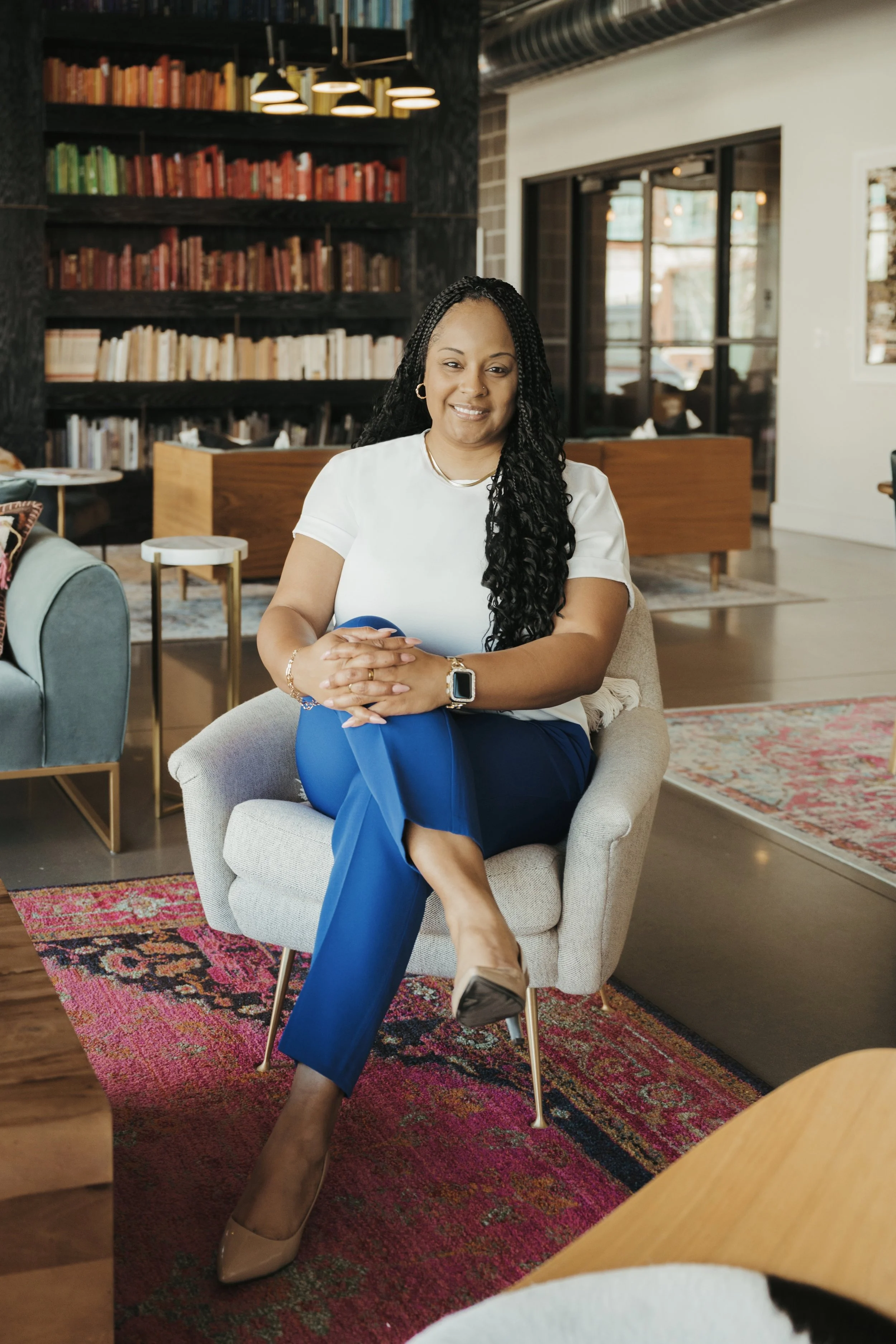 A woman with long braided hair, wearing a white t-shirt and blue pants, sitting on a beige armchair inside a modern library or bookstore with colorful bookshelves and large windows.