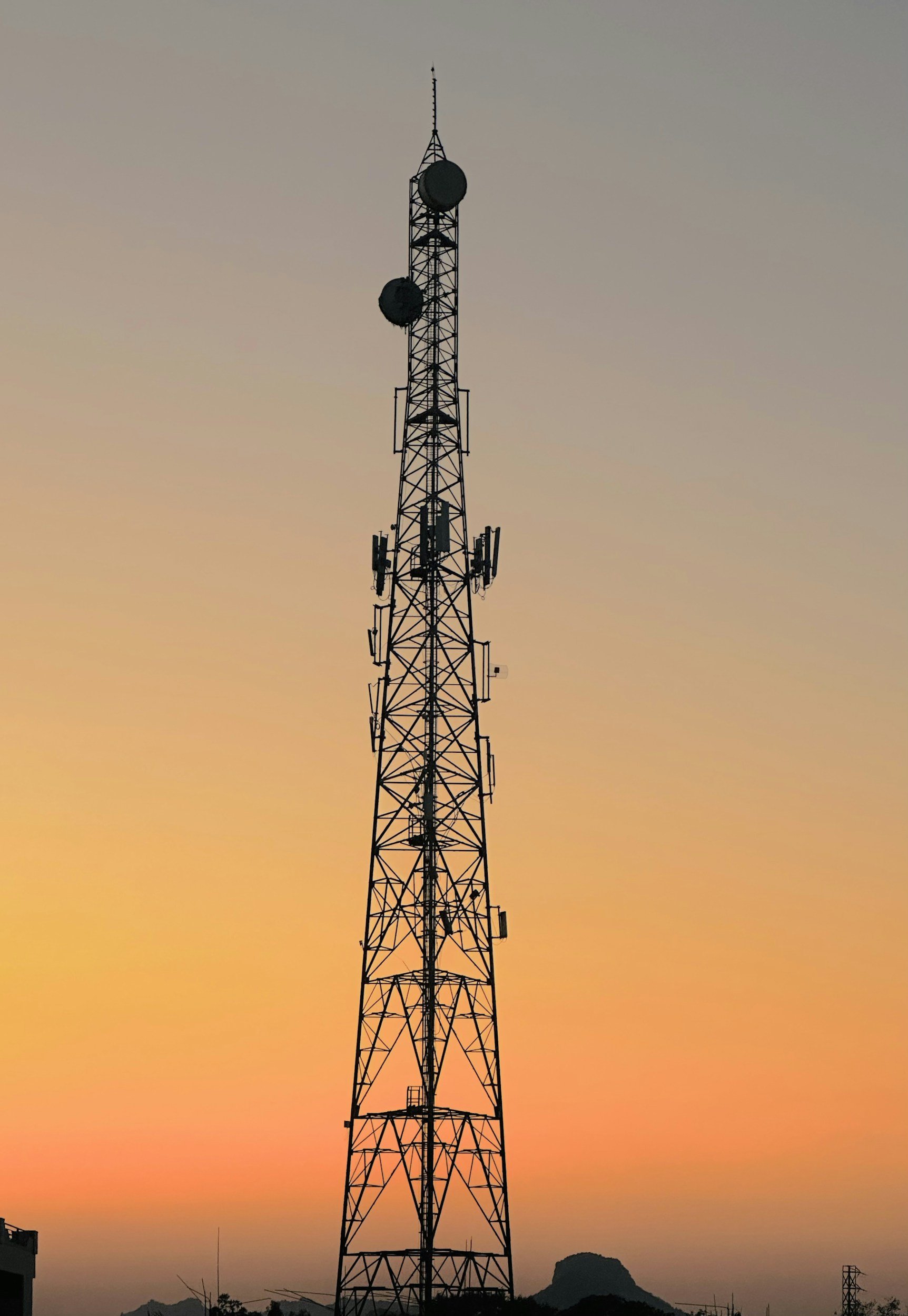 Tall radio tower with antennas and dishes against a sunset sky with mountains in the distance.