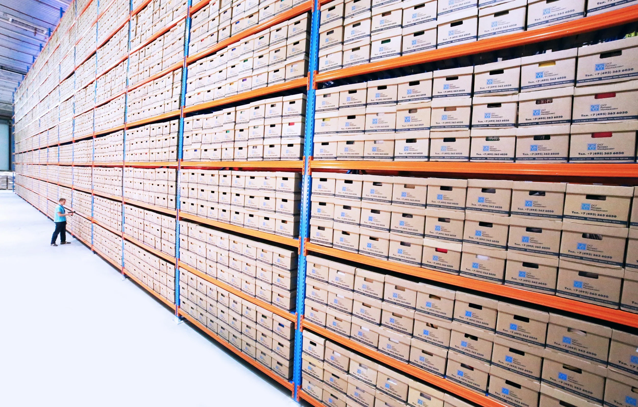 A large warehouse with tall shelves filled with packed cardboard boxes. A man in a blue shirt and black pants is standing near the shelves, looking at the boxes.