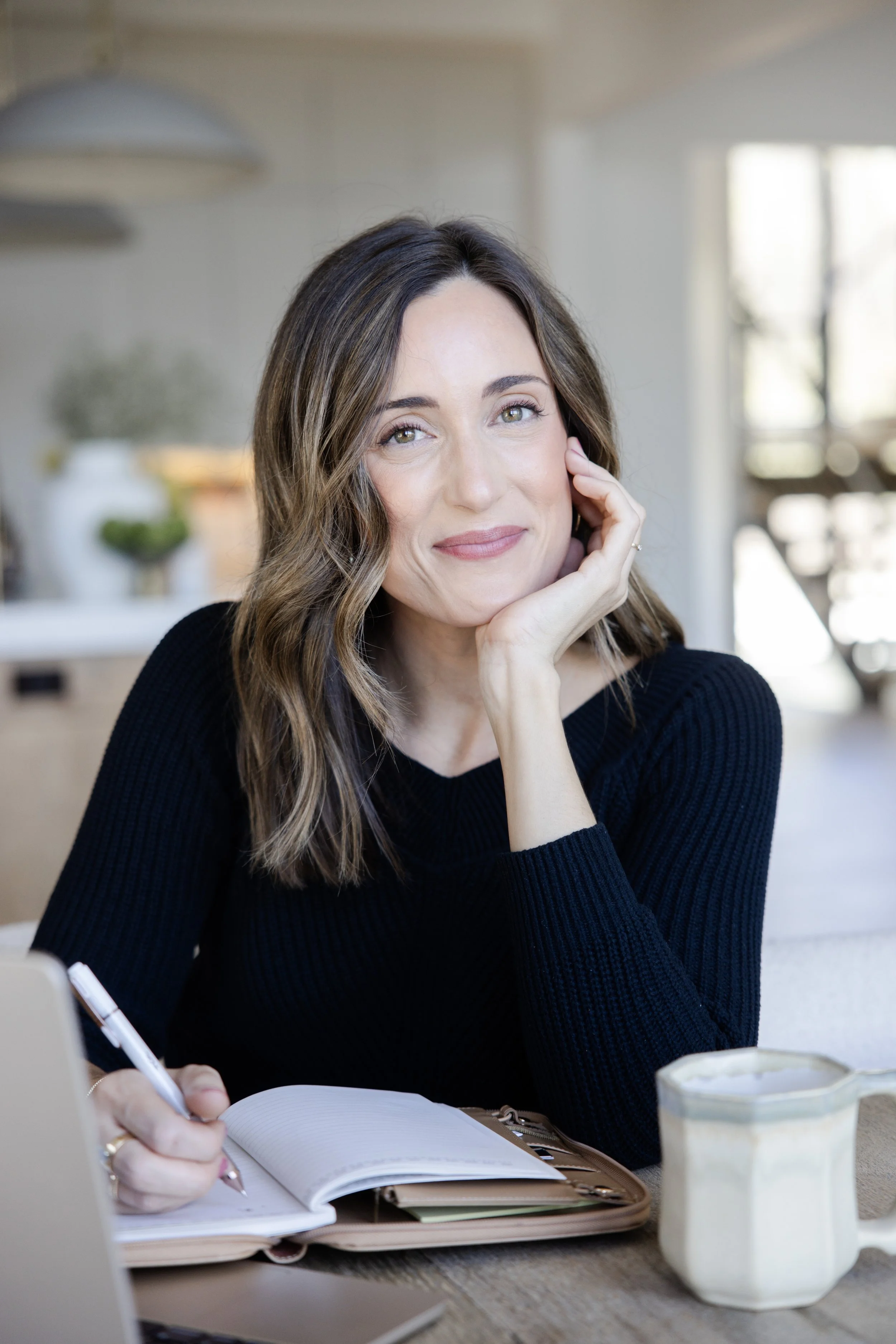 A woman with brown hair and light skin sitting at a desk, looking thoughtfully with her hand resting on her cheek. She has a notebook, a pen, and a laptop in front of her, with a large container of iced coffee nearby. The background shows a bright, modern kitchen.