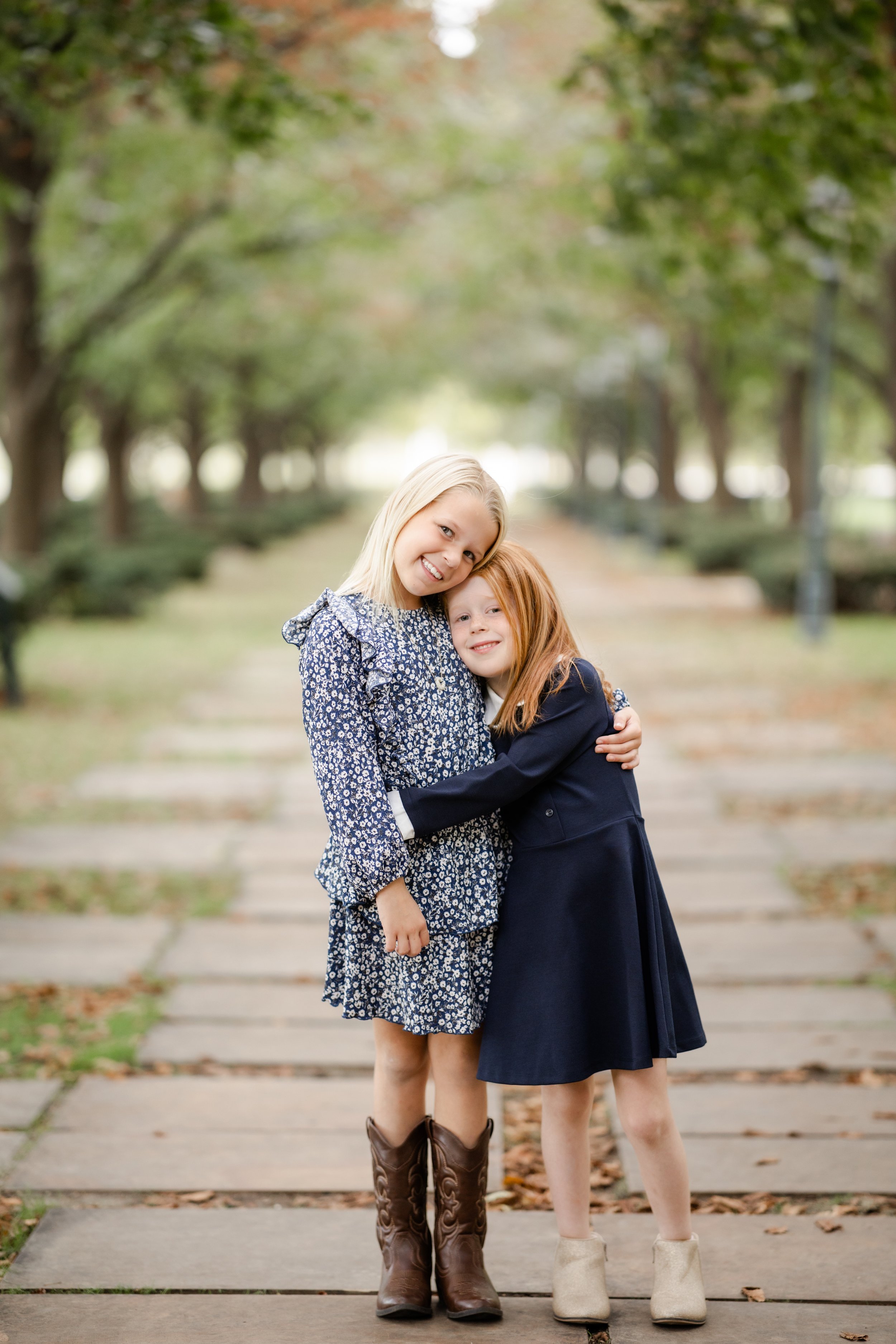 Two young girls with long hair, one with blonde hair and one with red hair, standing close together on a pathway in a park lined with trees, hugging and smiling at the camera.